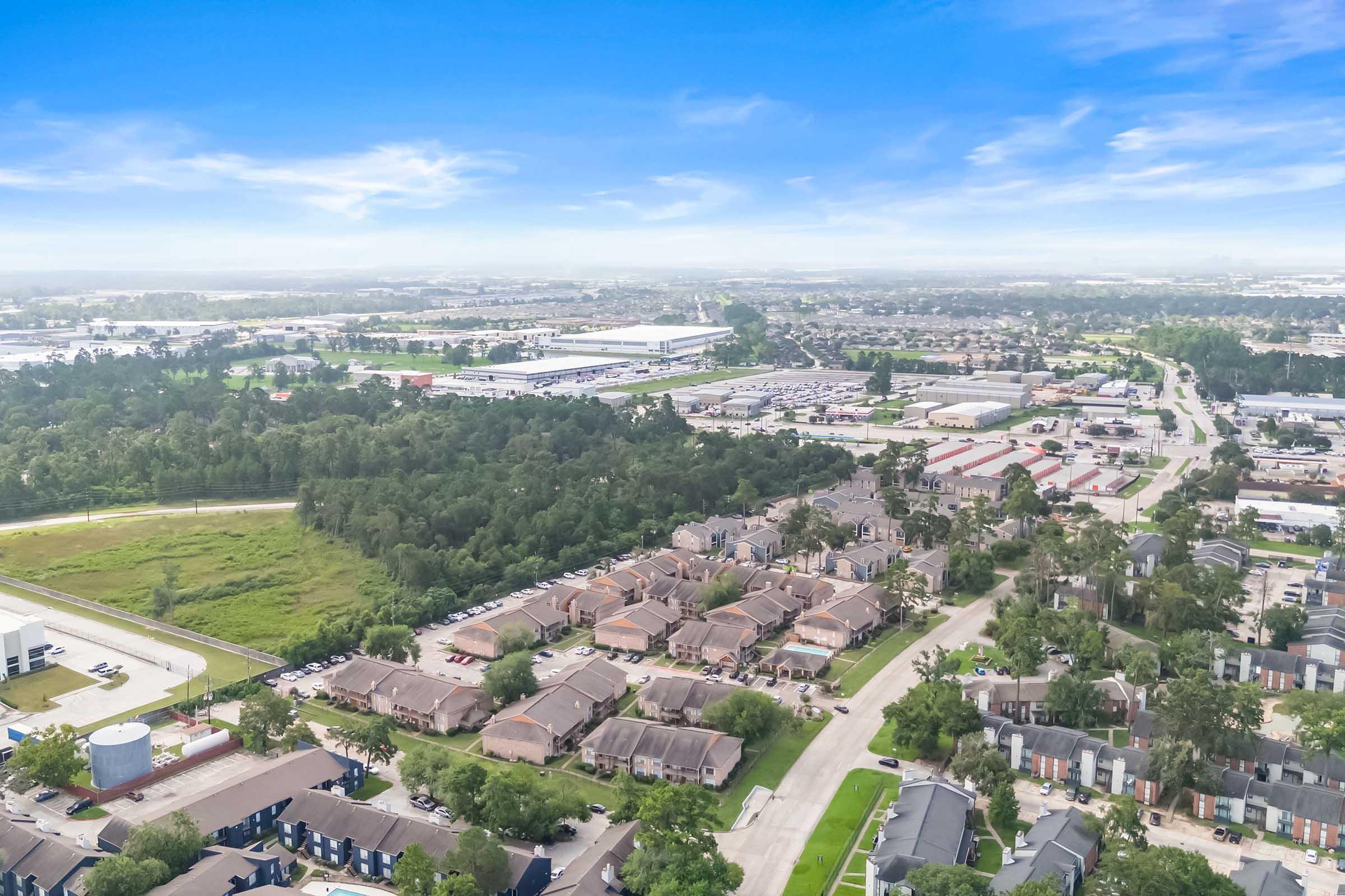 Aerial view of a suburban area featuring clusters of residential buildings, green spaces, and commercial facilities. The landscape includes a mix of housing, trees, and open fields with a clear blue sky above, showcasing a tranquil neighborhood setting.