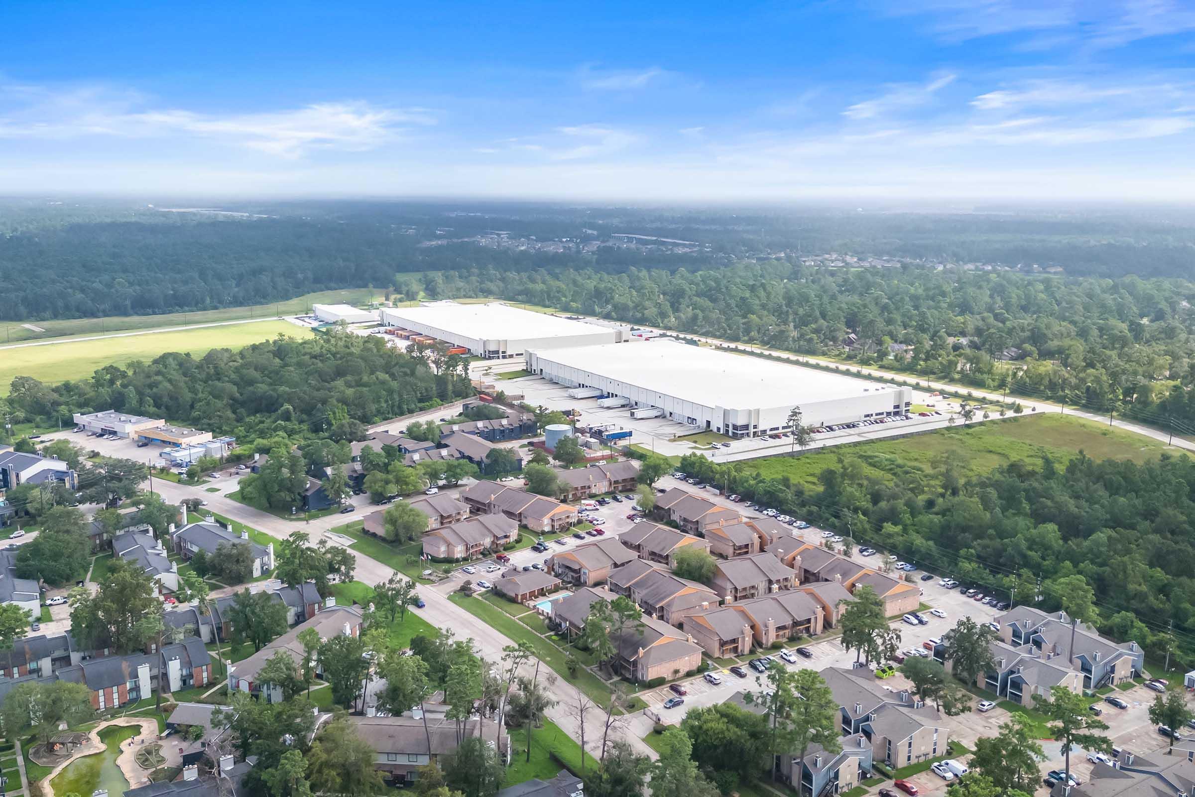 An aerial view of a large industrial complex with a white roof, surrounded by green trees and fields. In the foreground, there are residential buildings, likely apartments, with a parking area visible. The sky is clear with a few clouds, indicating a bright day.