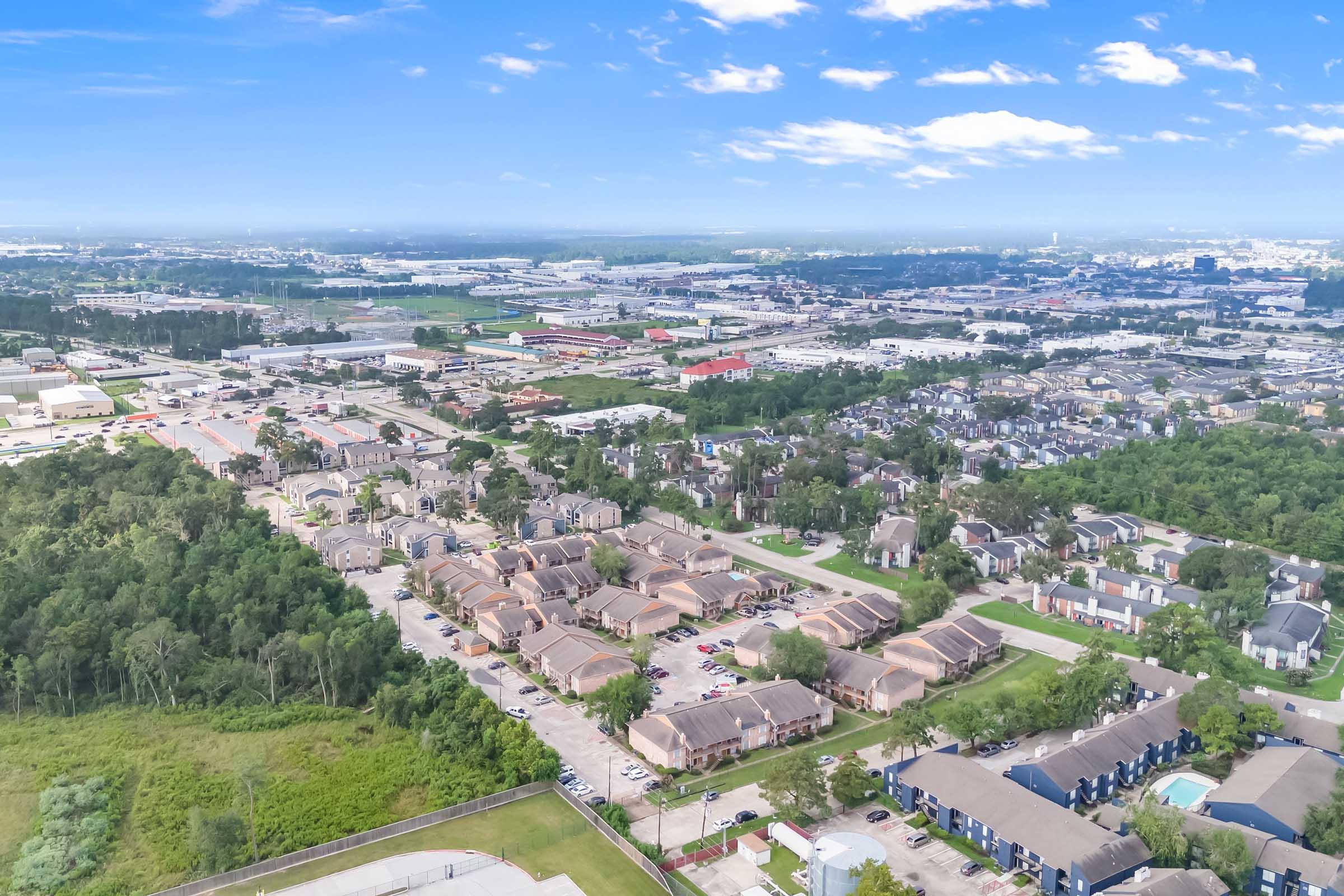 Aerial view of a suburban area featuring multiple residential buildings, green spaces, and a mix of urban development. The landscape includes a blend of houses and commercial properties, with clear blue skies and some clouds visible above.