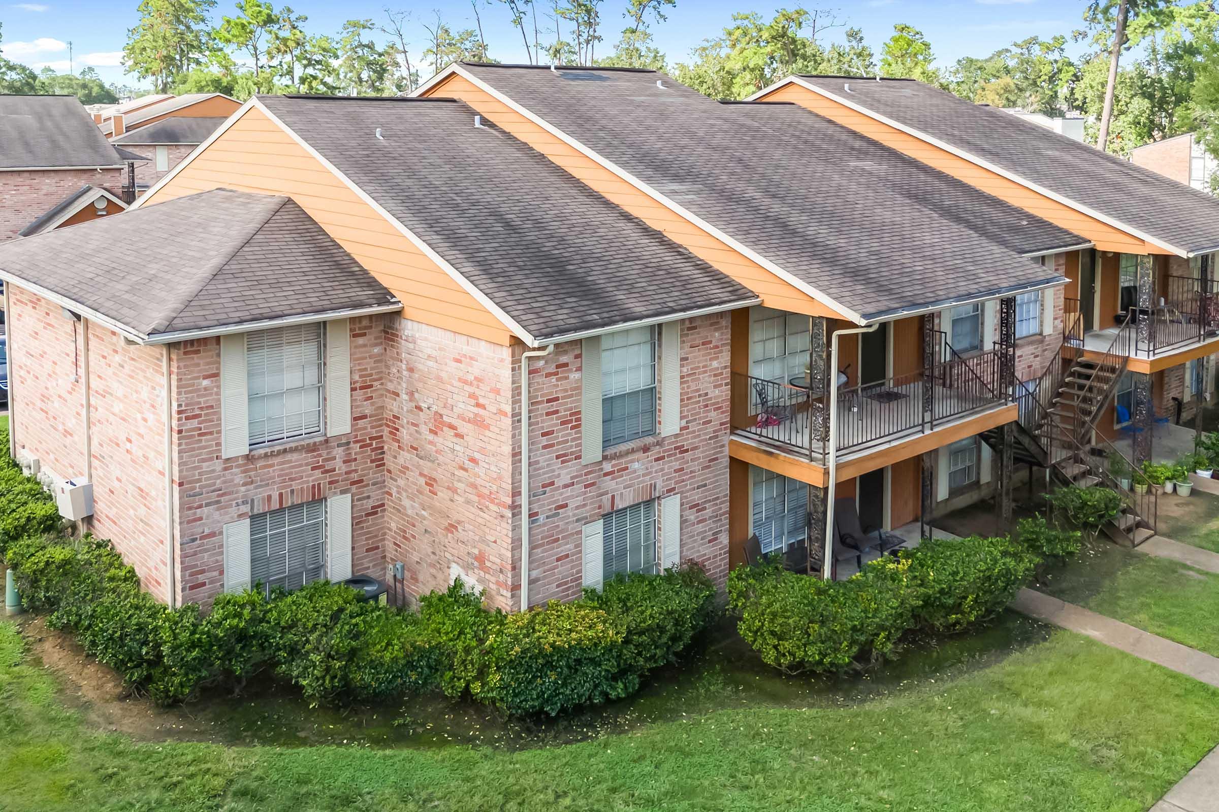 Aerial view of a residential apartment complex featuring brick buildings with orange accents, balconies, and a well-maintained lawn with shrubs. The layout includes multiple units and pathways, set in a green environment with trees in the background.