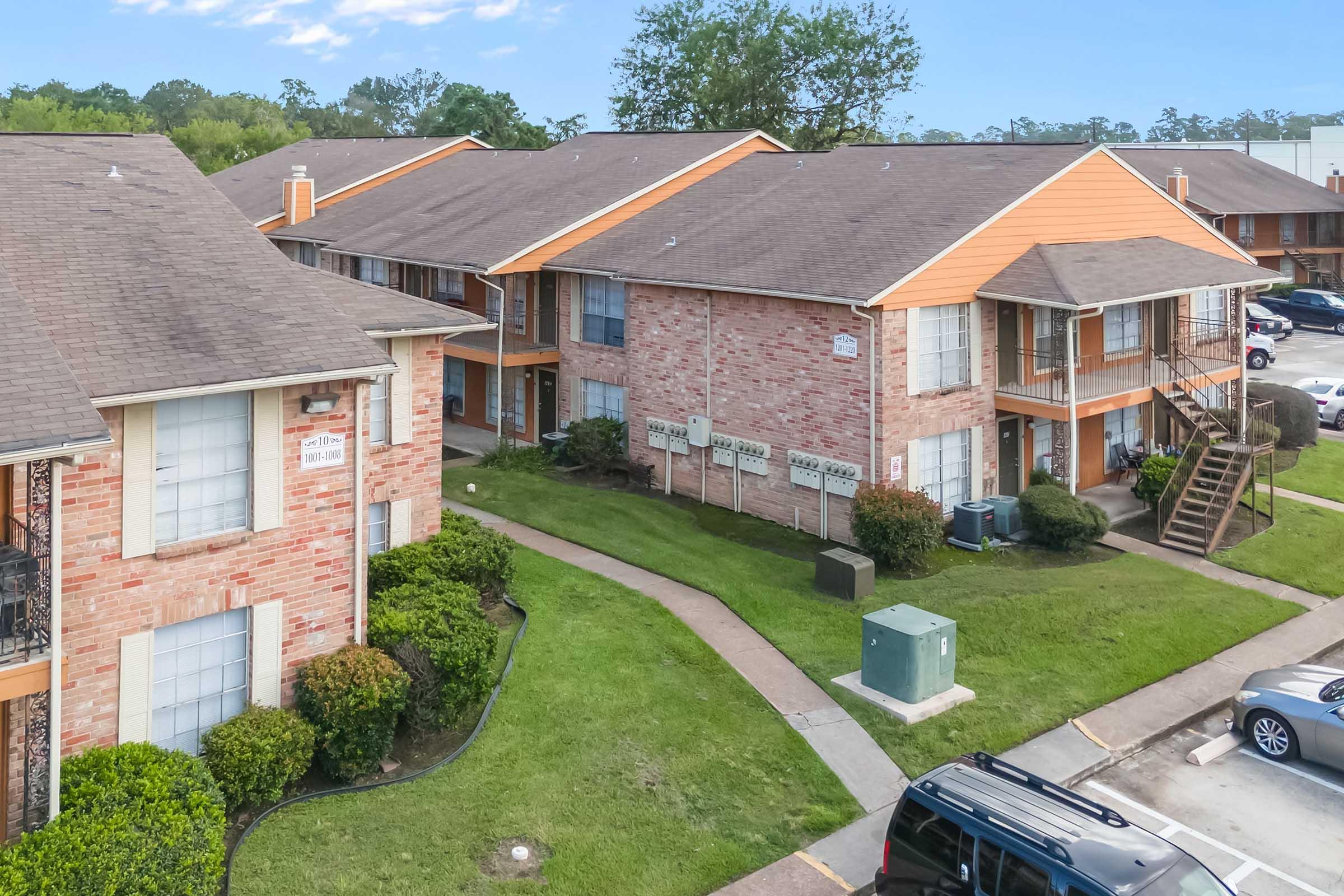 Aerial view of a residential complex featuring two-story brick apartments with orange accents, surrounded by green lawns and neat landscaping. The image shows pathways leading to individual units and parked cars nearby, set against a clear blue sky.