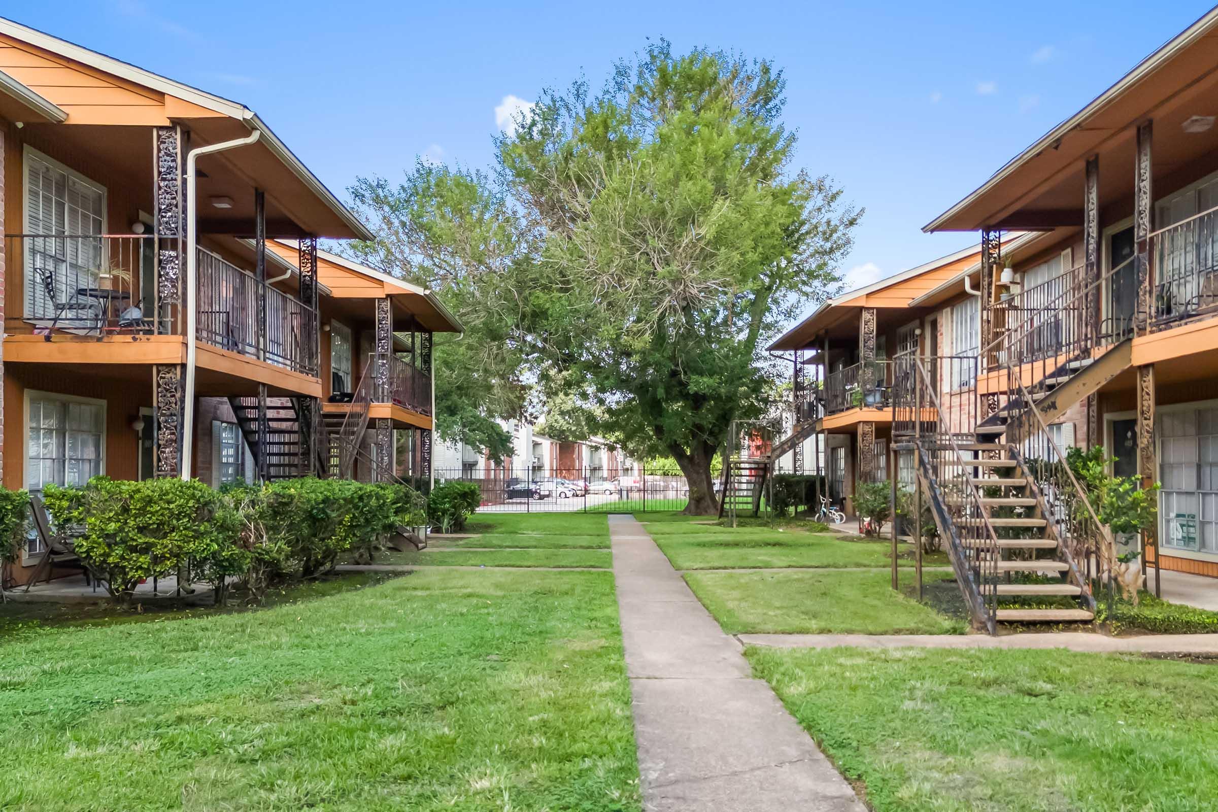 Two-story apartment buildings arranged around a central green lawn area. There are pathways leading through the grass, and each building features balconies with railings. Lush trees are present, adding greenery to the environment. The sky is blue with a few clouds, indicating a clear day.