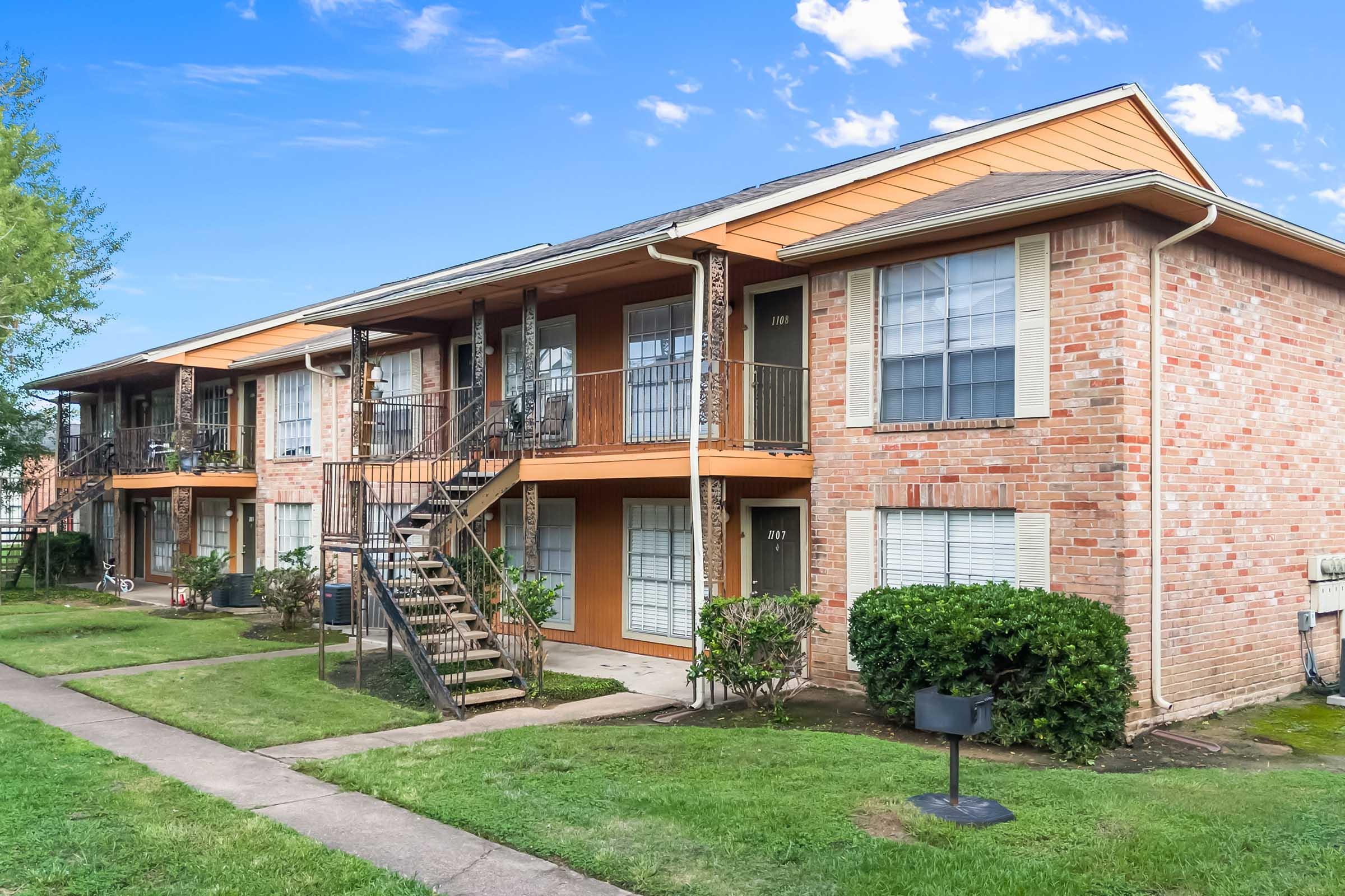 A two-story brick apartment building with orange accents, featuring multiple balconies and staircases. Green grass and bushes surround the building, with a concrete pathway leading to the entrance. The sky is clear with scattered clouds, creating a bright and inviting atmosphere.