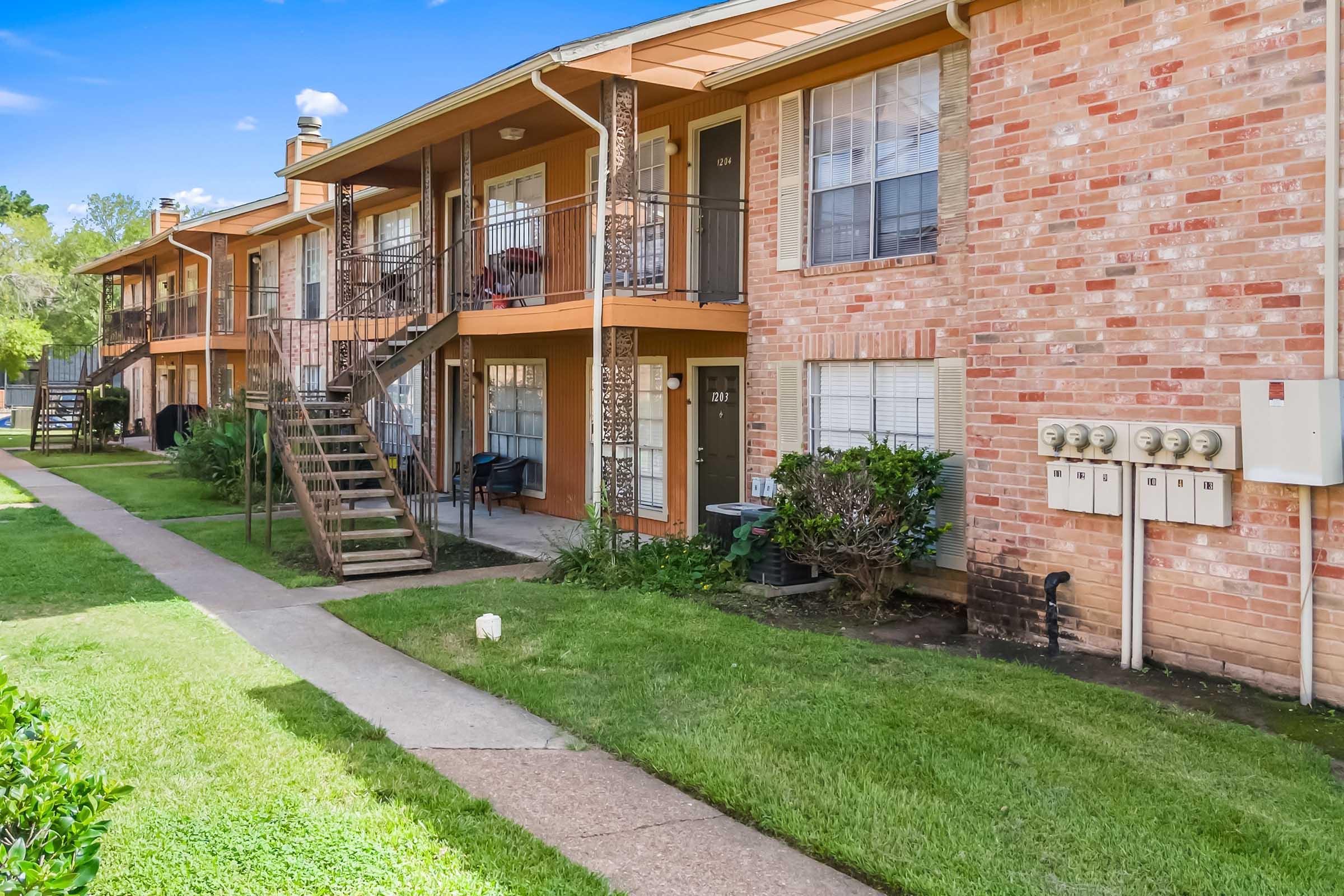 A view of a brick apartment building with two stories, featuring a well-maintained lawn and sidewalks. Staircases lead to the upper units, and there are utility meters on the side. Green shrubs and a white lawn ornament add to the outdoor appearance. Blue sky overhead.