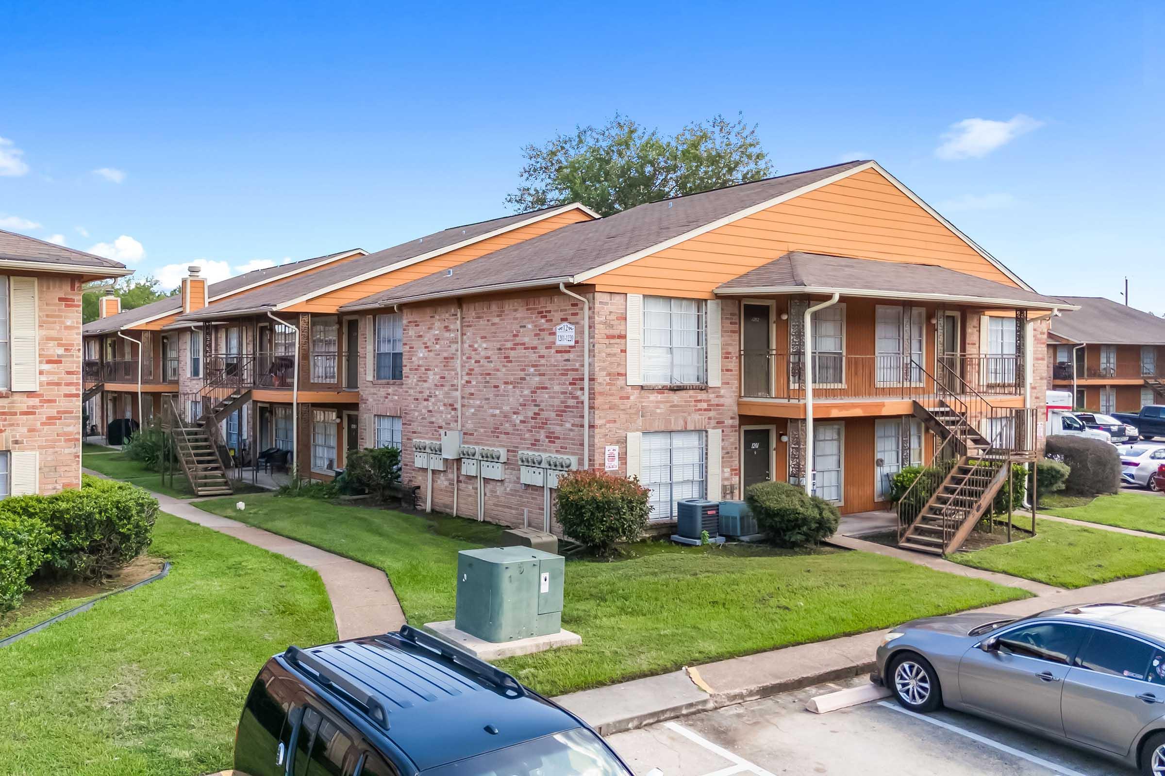 A multi-unit residential building with a brick exterior and orange accents. The structure features multiple entrances, staircases, and a well-maintained lawn. Nearby are parked cars and utility boxes, indicating a suburban setting. Clear blue skies provide a bright backdrop.