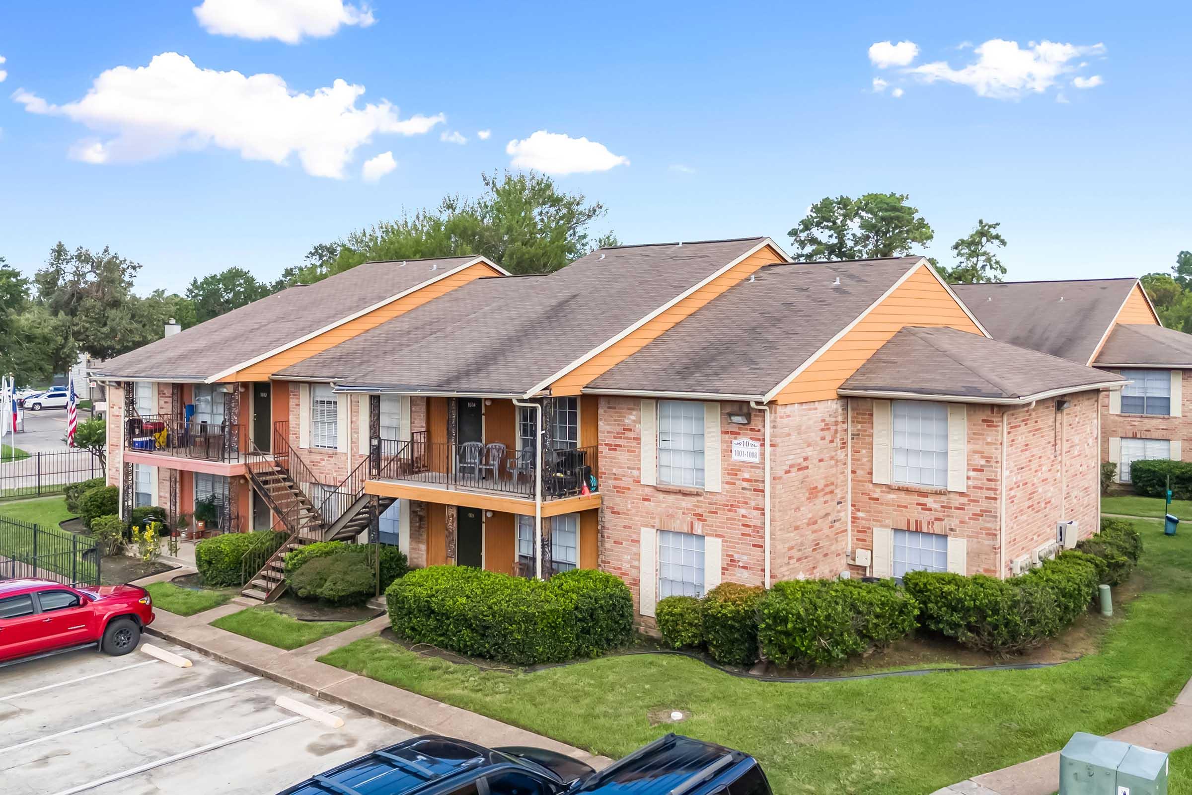 A view of a two-story apartment building with brick exterior and orange accents, surrounded by well-maintained greenery. The scene features parked cars in a lot, and a clear blue sky with a few clouds overhead.