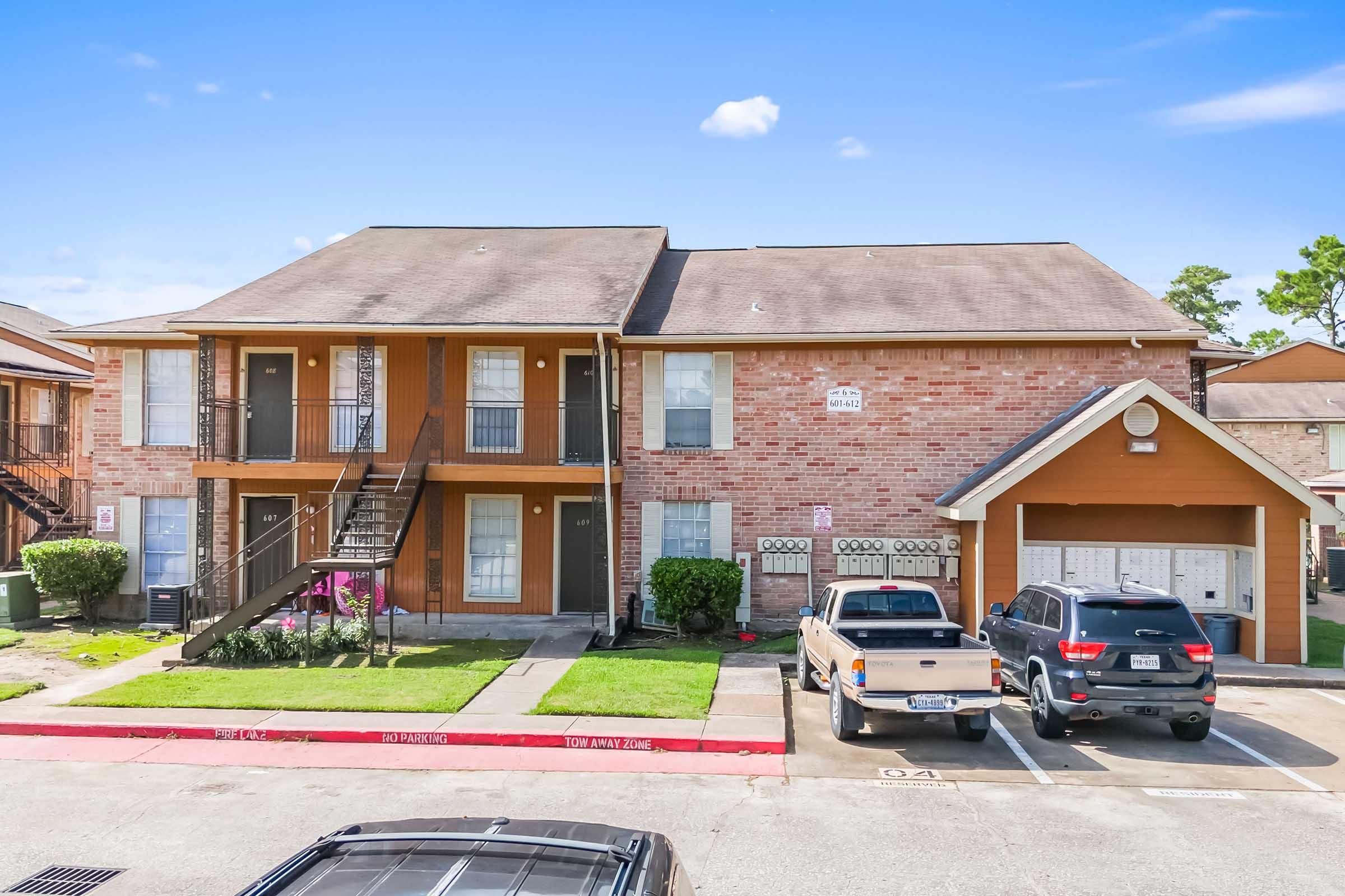 A two-story brick apartment complex with a parking lot. The building features balconies and a staircase leading to the upper level. There are two parked vehicles in front, one in a garage, and a landscaped area with flowers. The clear blue sky is visible in the background.