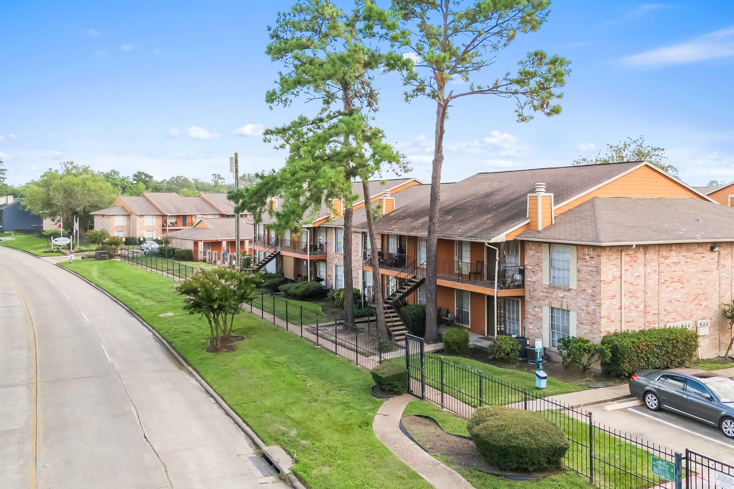 A view of a residential apartment complex with brick buildings and orange accents, surrounded by green lawns and trees. A paved road runs alongside the property, and several parked cars are visible. The scene is bright and clear, showcasing a peaceful neighborhood atmosphere.