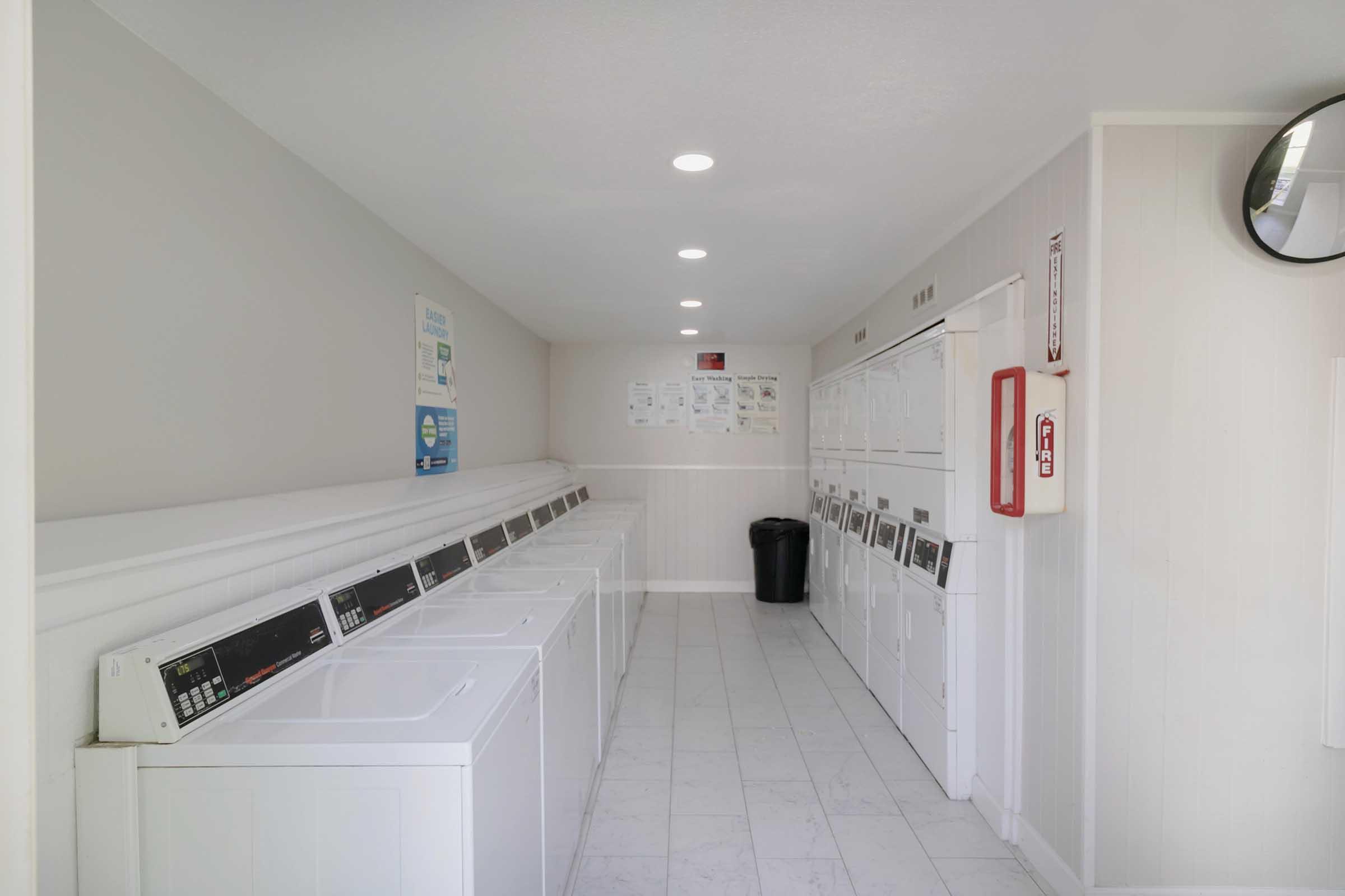A clean, well-lit laundry room featuring multiple washing machines and dryers lined up against the wall. There are white cabinets with notices posted above, a black trash bin in the corner, and a mirror on the wall reflecting the space. The flooring is tiled in light colors.