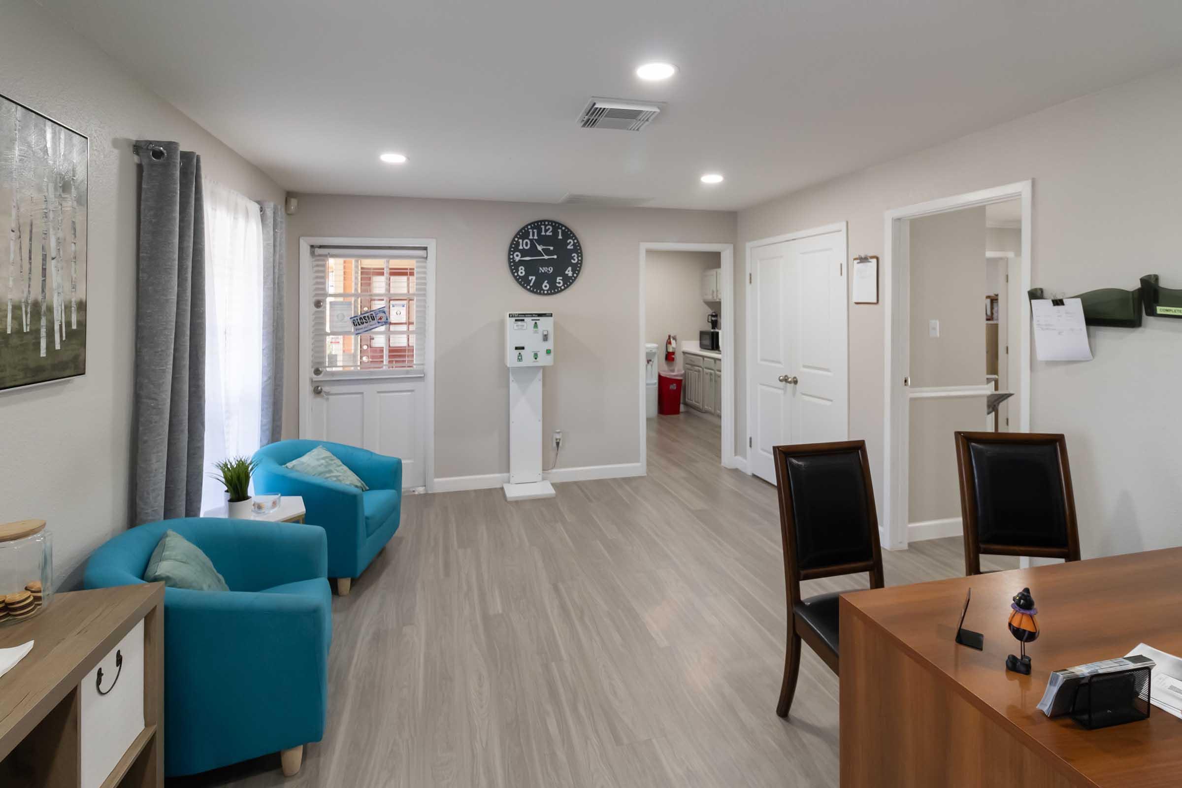 A bright, modern office space featuring two blue chairs, a wooden desk with a decorative item, and a large wall clock. The walls are painted light colors, and there is a window with sheer curtains letting in natural light. A hallway leads to another room, visible in the background.