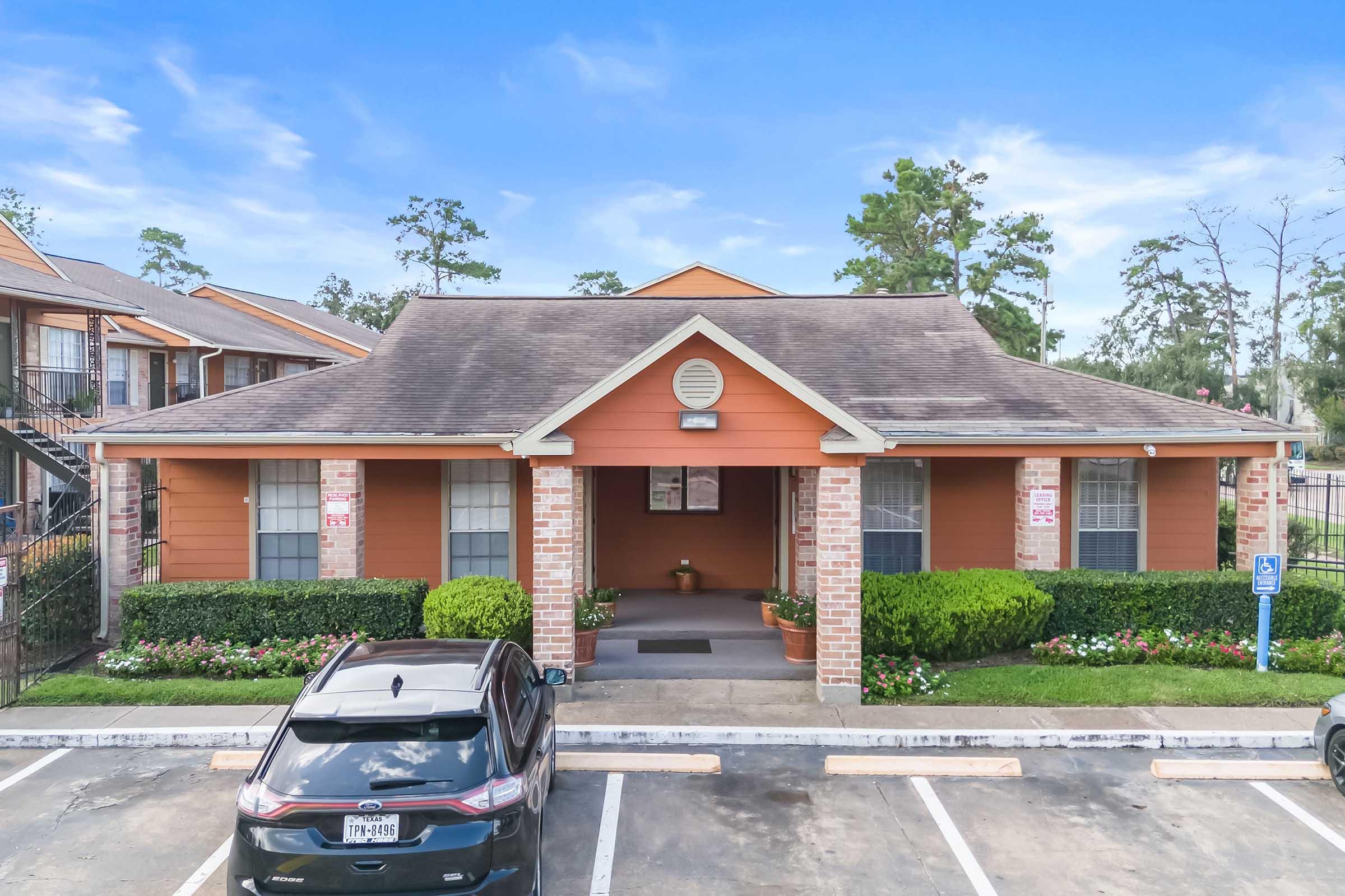 Front view of a single-story building with orange siding and a peaked roof. The entrance is framed by brick pillars and surrounded by neatly trimmed hedges and potted plants. Several parking spaces are visible in front, with a car parked nearby. Trees and a blue sky provide a pleasant backdrop.