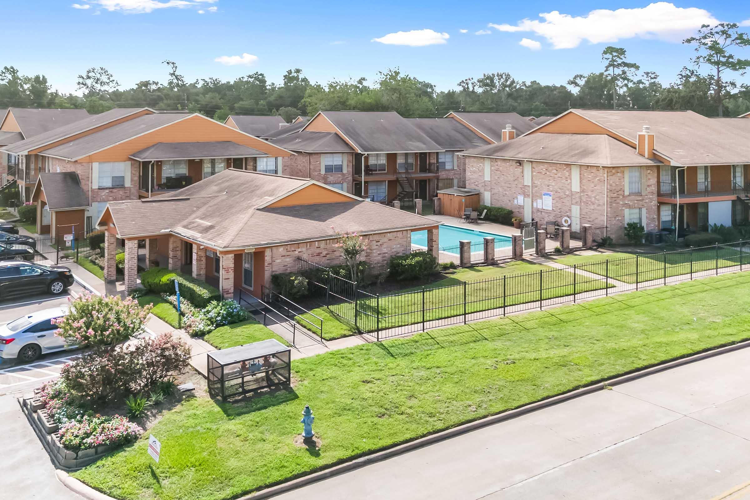 Aerial view of a residential complex featuring multiple two-story buildings with orange roofs, a swimming pool, neatly landscaped lawns, and parking spaces. Surrounding greenery adds to the tranquil atmosphere. Clear blue sky in the background enhances the appeal of the property.