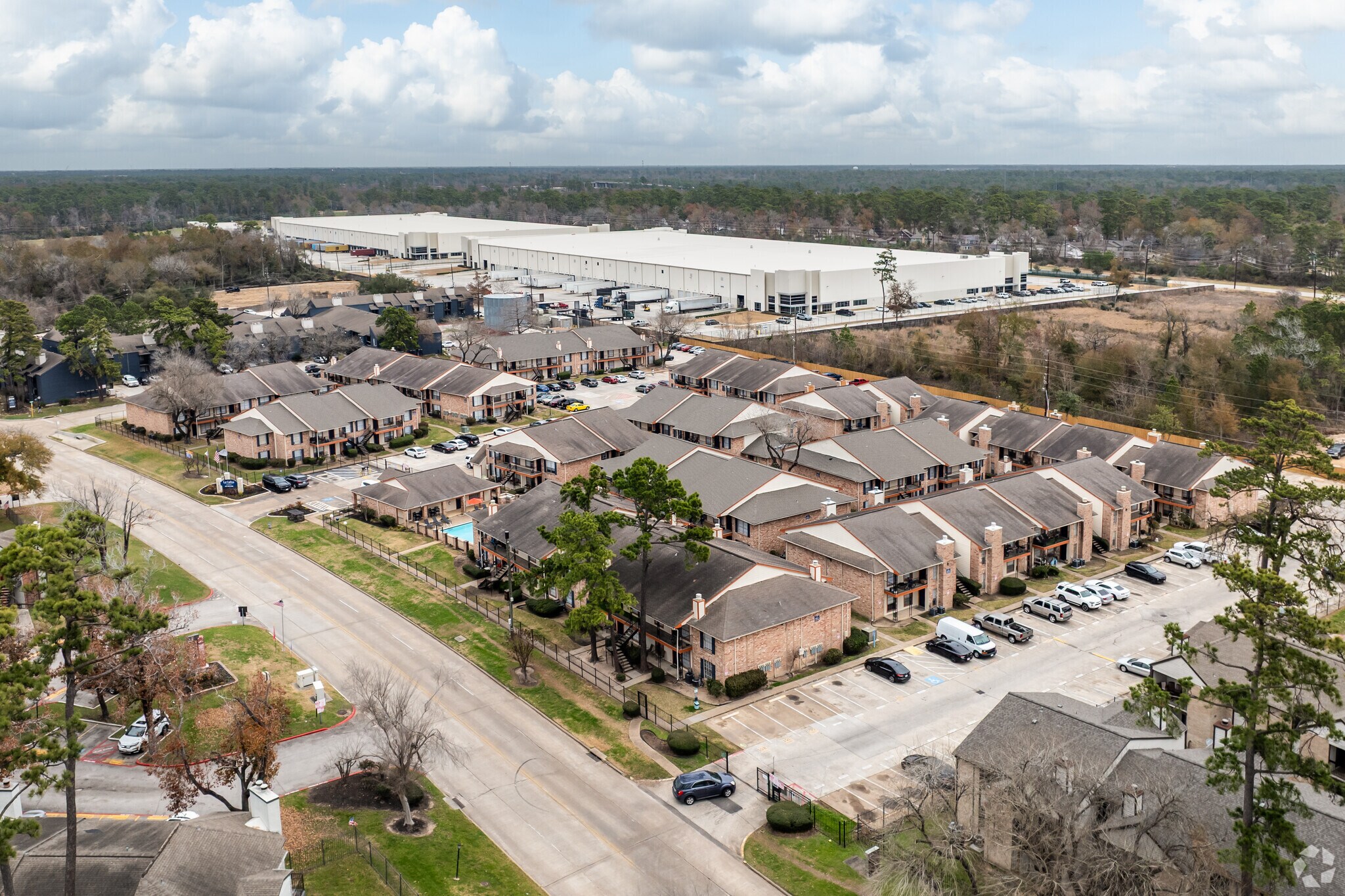 Aerial view of a residential area featuring several two-story apartment buildings with parking lots, surrounded by trees. In the background, a large industrial warehouse building is visible, set against a partly cloudy sky. The scene exhibits a suburban landscape with a mix of housing and commercial structures.