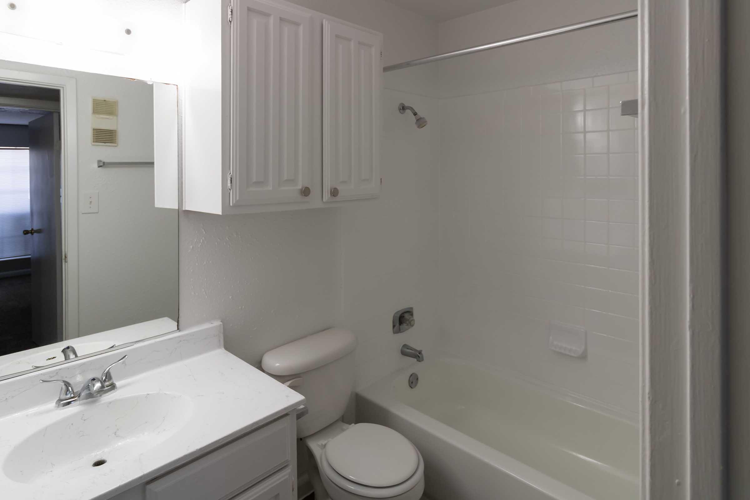 A clean, simple bathroom featuring a white bathtub with a showerhead, a sink with a mirror, and a white toilet. The walls are light-colored, and there are white cabinets above the sink for storage. A doorway leads to another room, partially visible in the background.