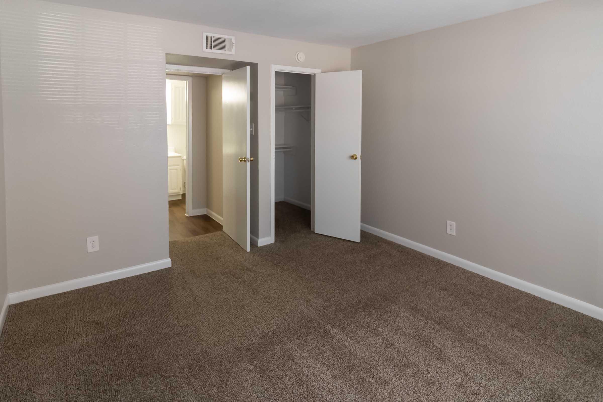 A well-lit room with beige walls and carpet flooring. The room features two open doors leading to a closet and a hallway. Natural light filters in, creating a spacious and inviting atmosphere. The layout is simple and modern, suitable for various uses.