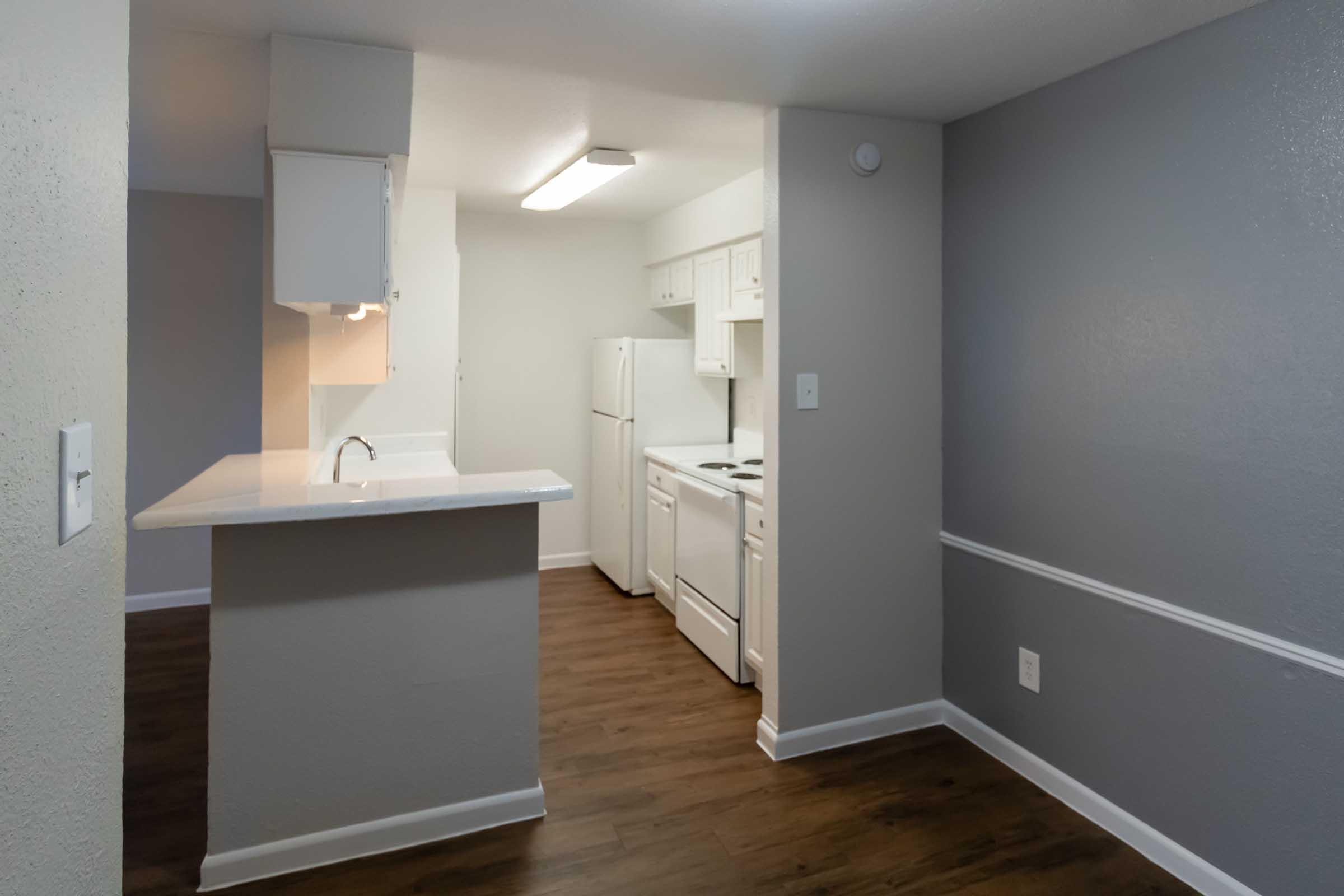 Modern kitchen with white cabinetry and appliances, including a refrigerator and stove. The open layout features a bar counter separating the kitchen from the living area. Neutral gray walls and wood-like flooring create a contemporary, inviting atmosphere.