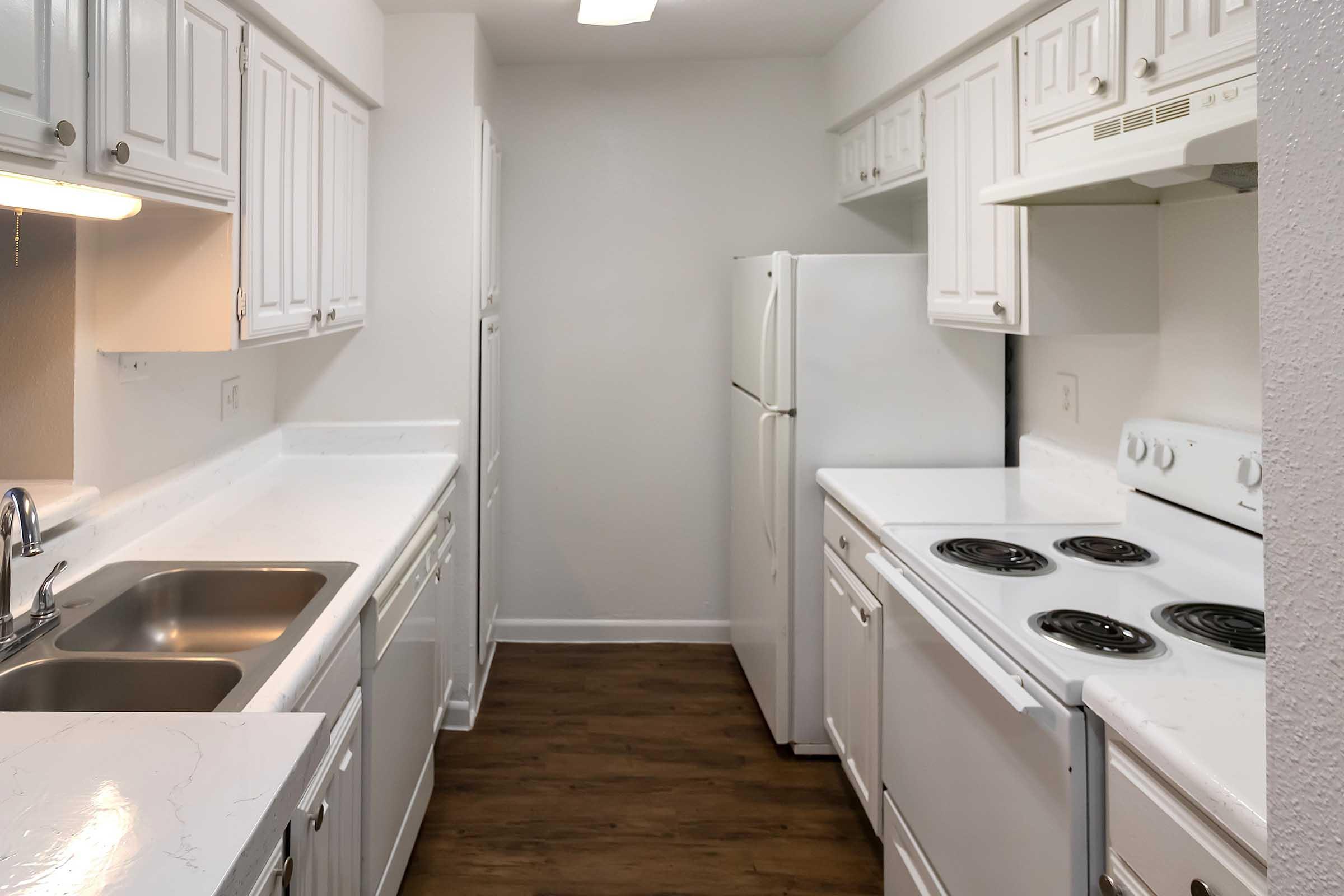 Bright, modern kitchen featuring white cabinetry, a double sink, and appliances including a refrigerator and stove. The countertop is clean and uncluttered, with a light color scheme and wood-style flooring, creating a spacious and inviting atmosphere.