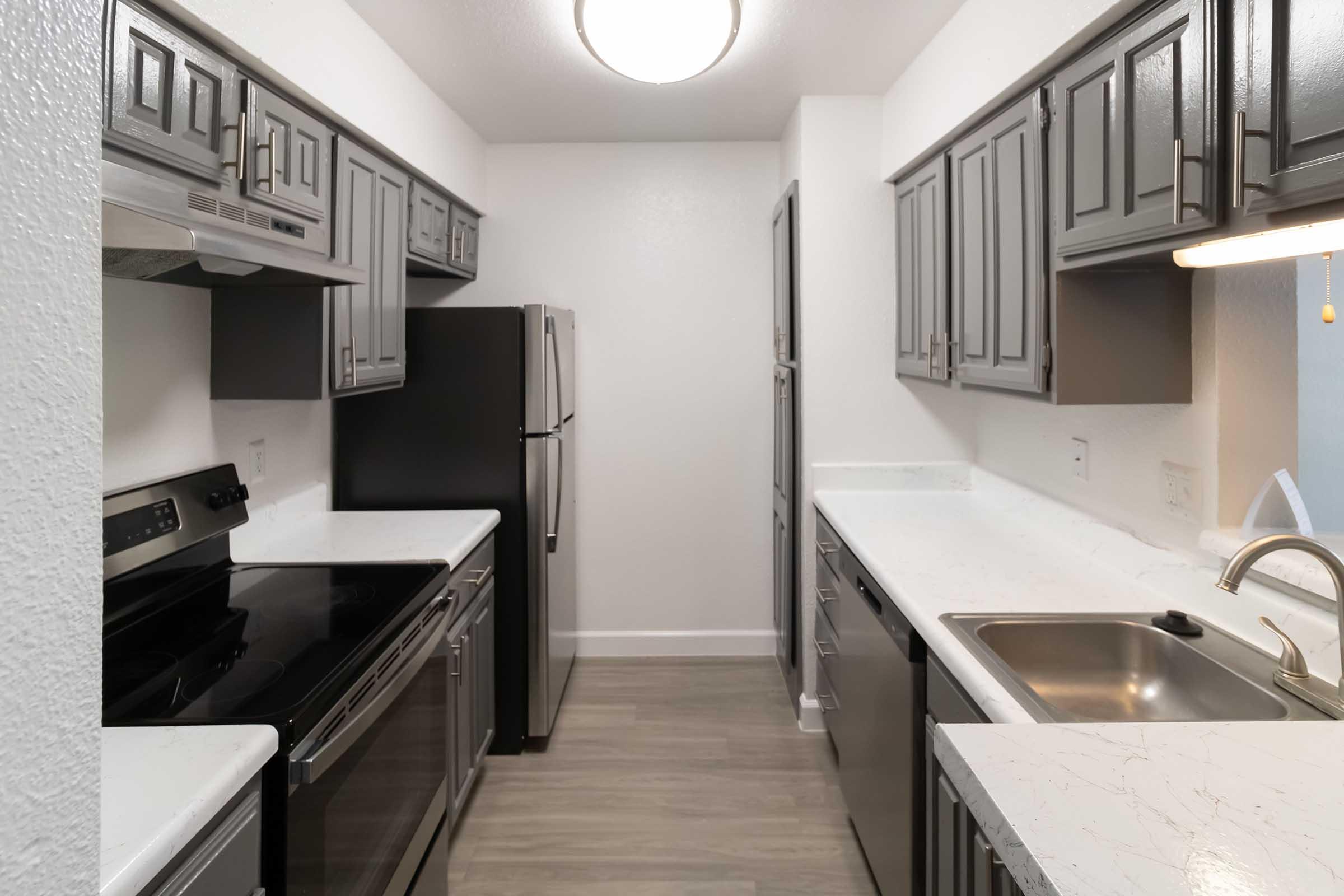 Modern kitchen featuring gray cabinetry, stainless steel appliances including a refrigerator, oven, and dishwasher, and a white countertop. The space has a clean, minimalist design with neutral tones and ample lighting.