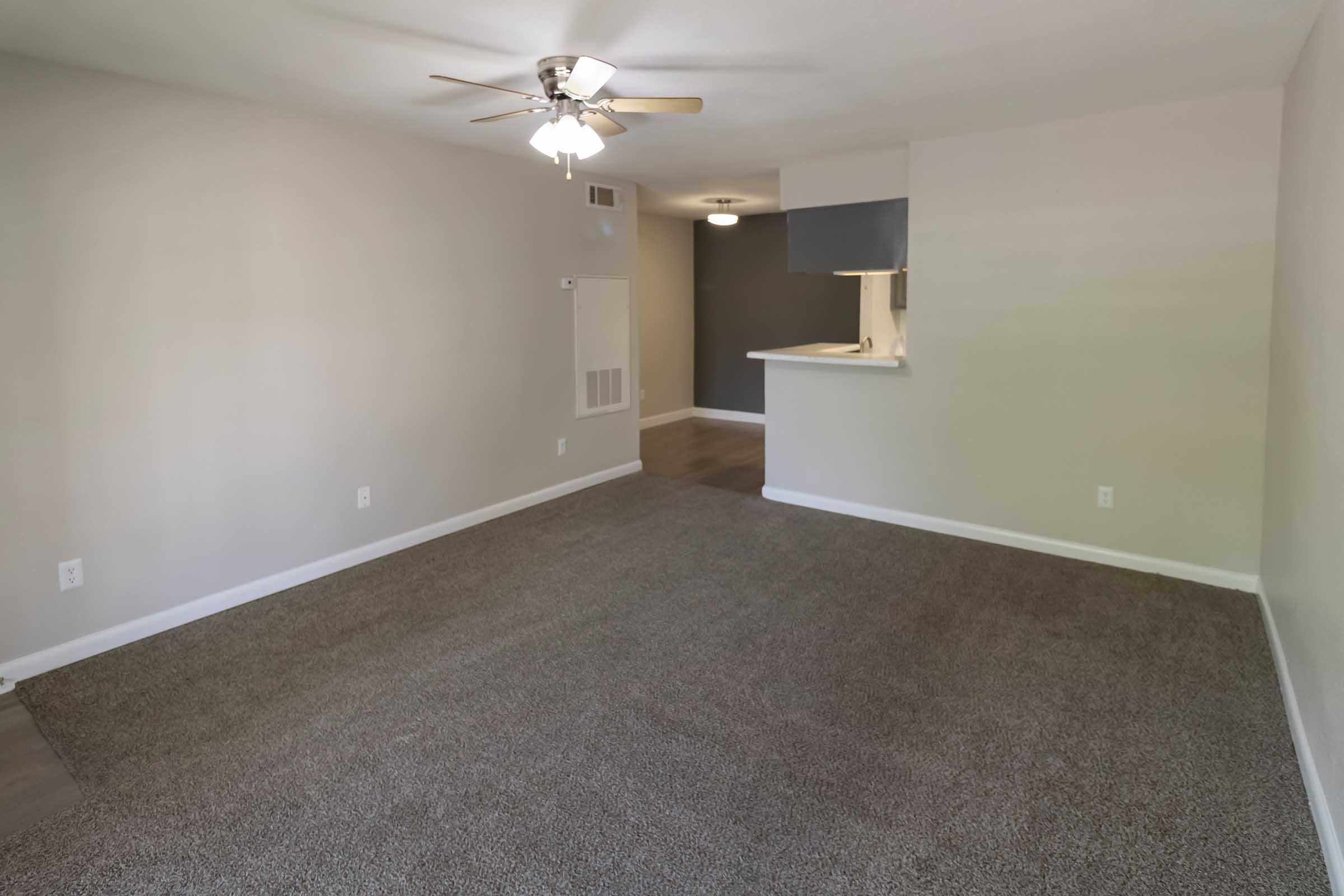 A spacious, unfurnished living room with beige walls and light gray carpeting. A ceiling fan is installed, and a doorway leads to a kitchen area with a bar counter. The room has ample natural light and a neutral color scheme, creating a bright and inviting atmosphere.