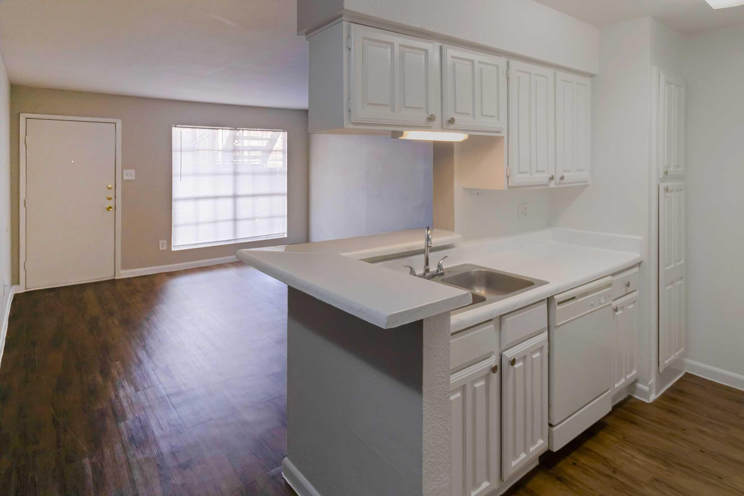 A modern kitchen with white cabinets and appliances, featuring a sink and dishwasher, seamlessly connected to a spacious living area with hardwood flooring and a large window allowing natural light. The entrance door is visible, creating an open and airy atmosphere.