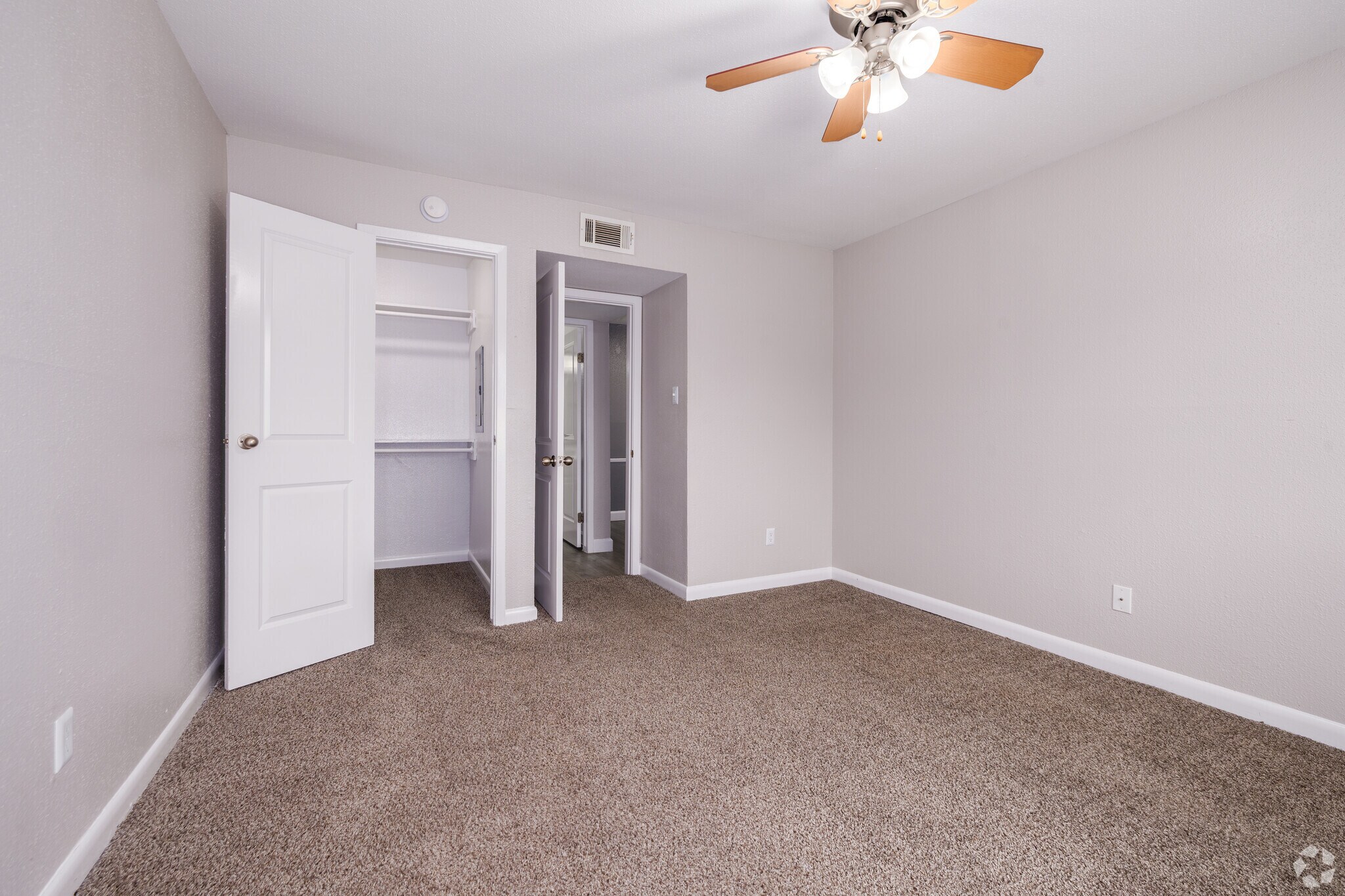 Empty room featuring light beige walls and plush carpet. A ceiling fan is visible, and there are two open doors leading to closets. The natural light brightens the space, creating an inviting atmosphere. The room is spacious and ready for furnishing.