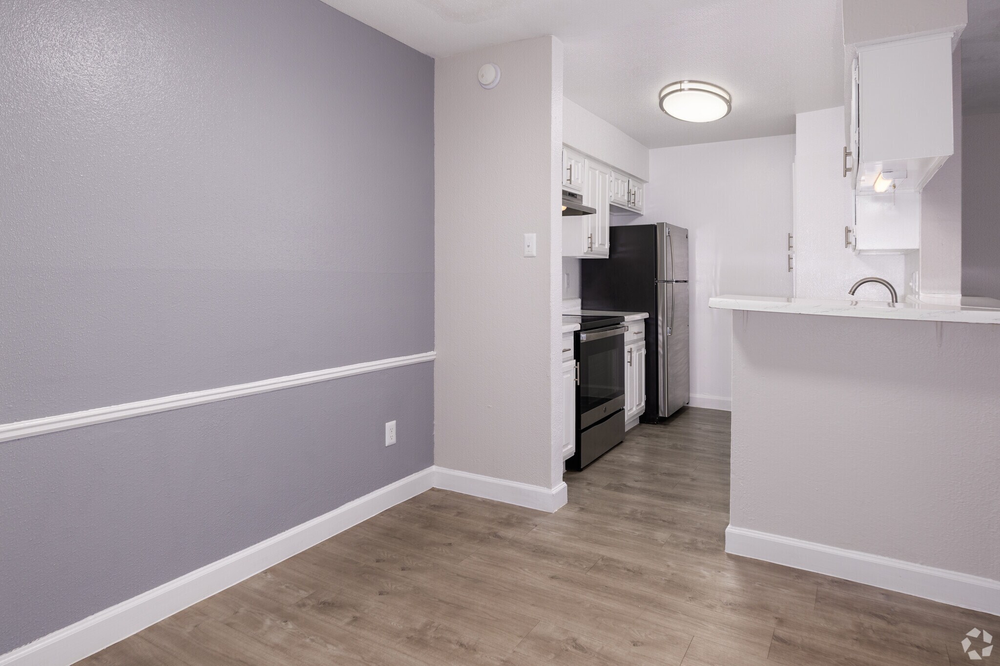 A modern kitchen featuring stainless steel appliances, including an oven, refrigerator, and dishwasher. The walls are painted in a light gray, with a darker accent wall. The floor is a light wood laminate, and the space is brightened by a ceiling light fixture. Counter space is available at the bar.