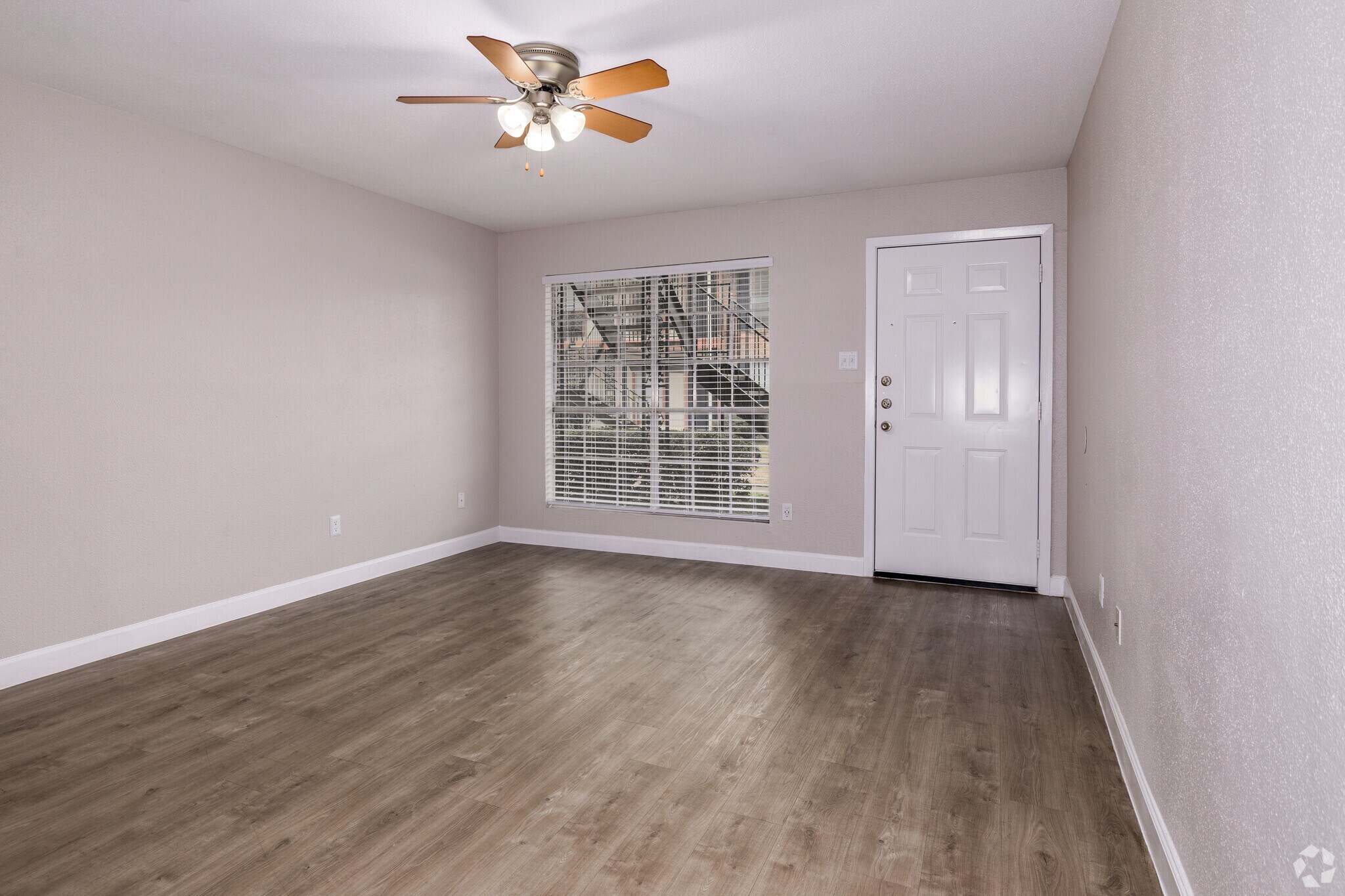 A well-lit empty room with a ceiling fan, light-colored walls, and laminate flooring. There is a window with horizontal blinds and a doorway leading outside. The space appears clean and uncluttered, suitable for various uses or furniture arrangements.