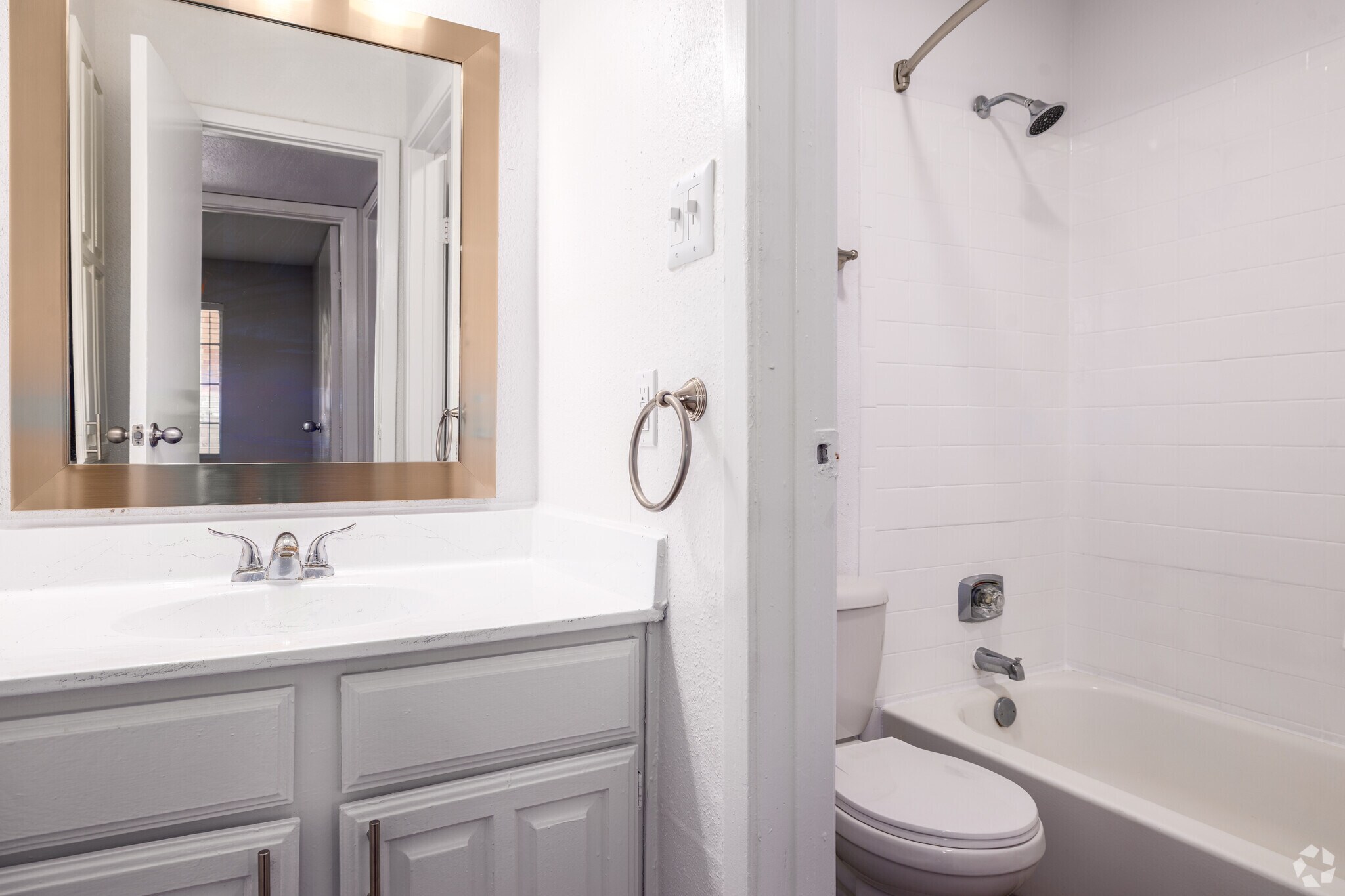 A clean, modern bathroom featuring a white vanity with a mirror, a sink, and a towel ring. There is a bathtub with a showerhead, a toilet, and light-colored walls. An open doorway leads to another room, creating a bright and spacious atmosphere.