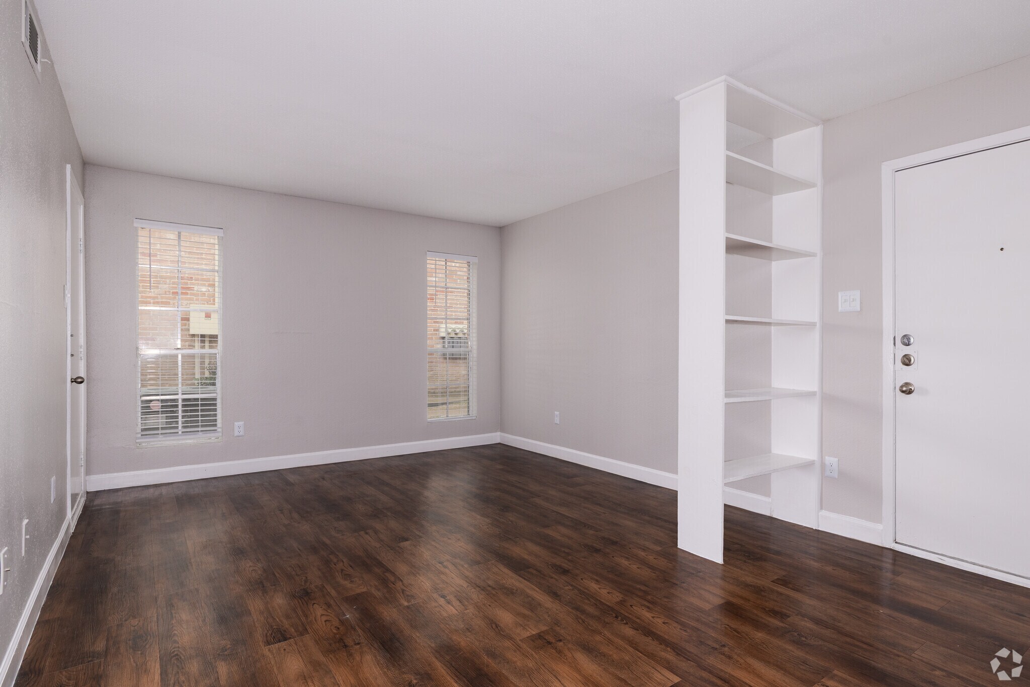An empty room with light gray walls and a dark wood floor. Two windows let in natural light, and there's a small white bookshelf against the wall. A door is visible on the right side of the image. The space appears spacious and well-lit, suitable for various interior design options.