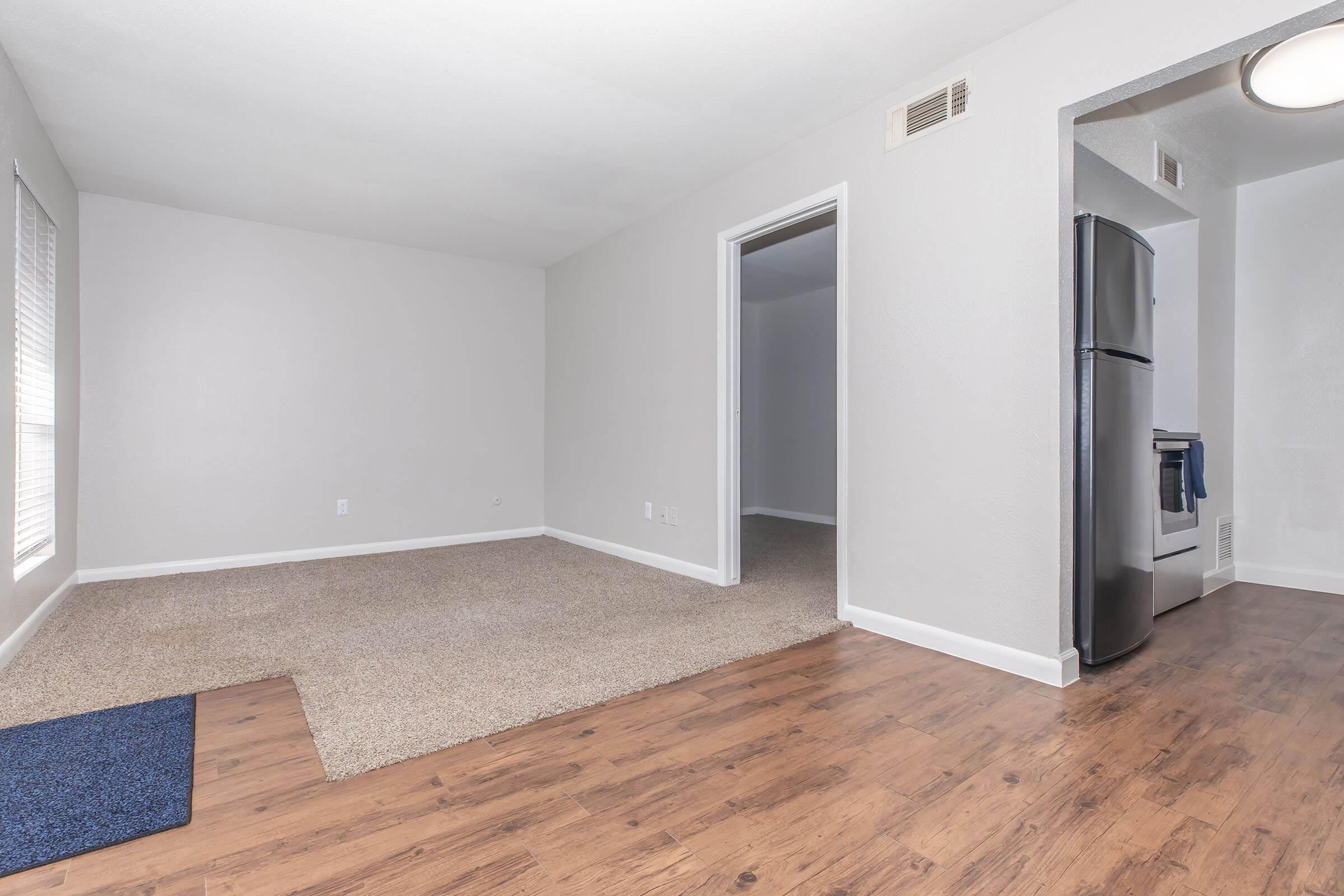 A spacious living area with light-colored walls and carpet flooring. There is a blue rug on the floor, a door leading to another room, and a glimpse of a kitchen area with a black refrigerator in the background. The space is well-lit with natural light coming through the windows.