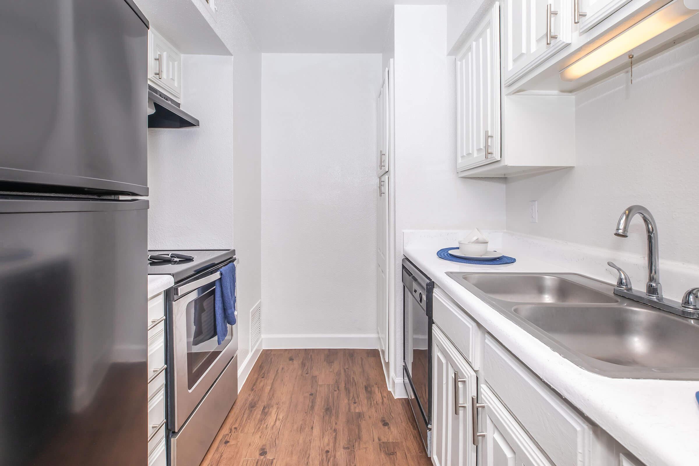 A modern kitchen with a refrigerator, stove, and sink. The cabinetry is white, and the countertops are light-colored. A dish is placed on a blue mat next to the sink. The flooring is wood-like, and the walls are painted in a light color, creating a bright and open atmosphere.