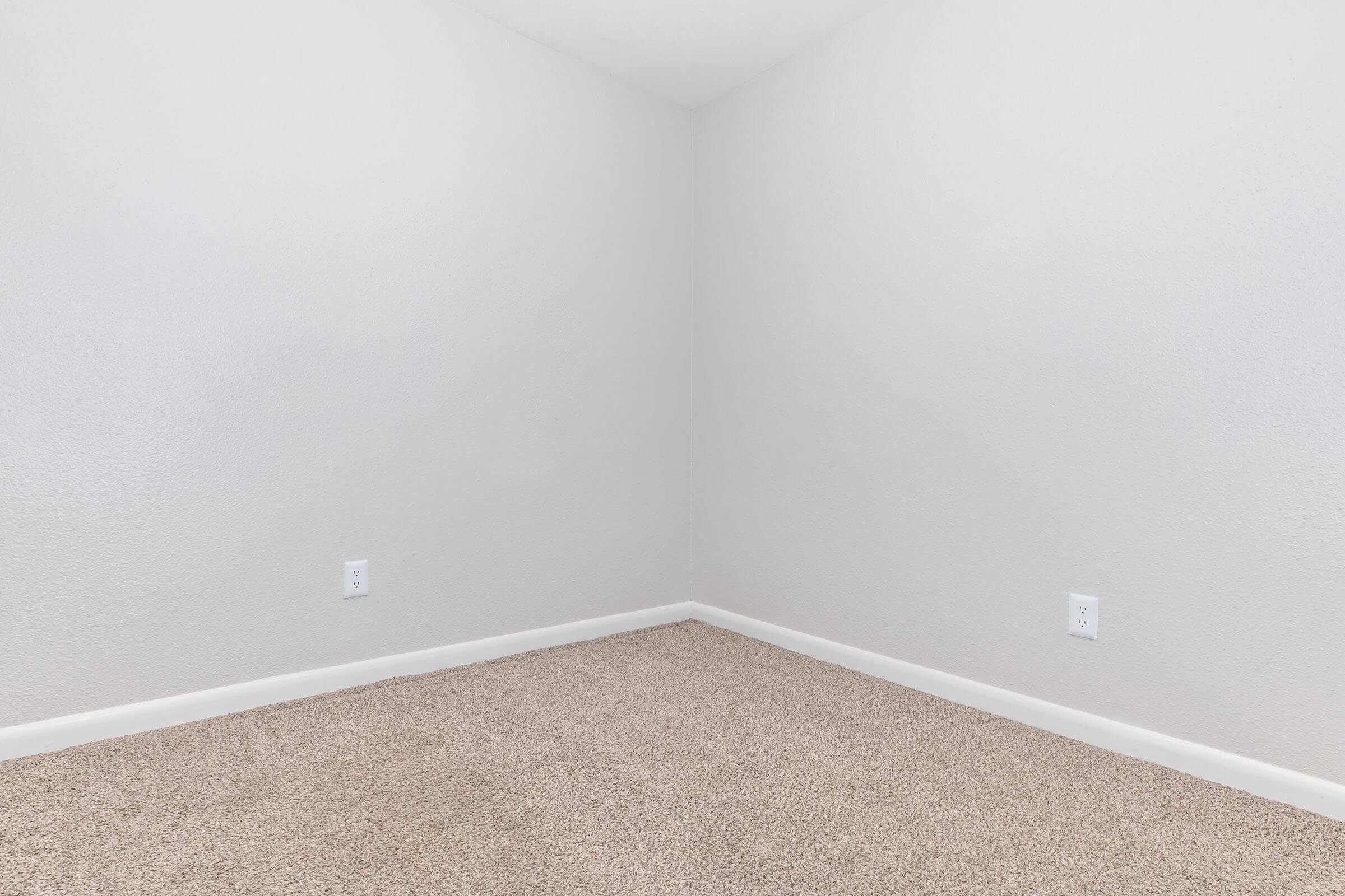 Empty room with light gray walls and a beige carpeted floor. The room features a corner where two walls meet, and there are no furniture or decorations visible. There are electrical outlets on the walls, and the space appears bright and clean.