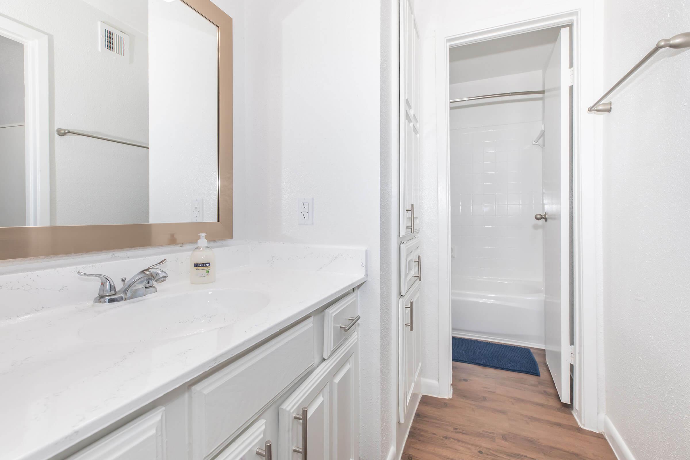 A modern bathroom featuring a white sink with a large mirror above, light-colored cabinetry, and a clean shower area visible through an open door. The walls are painted white, and there is a blue mat on the floor in front of the shower.