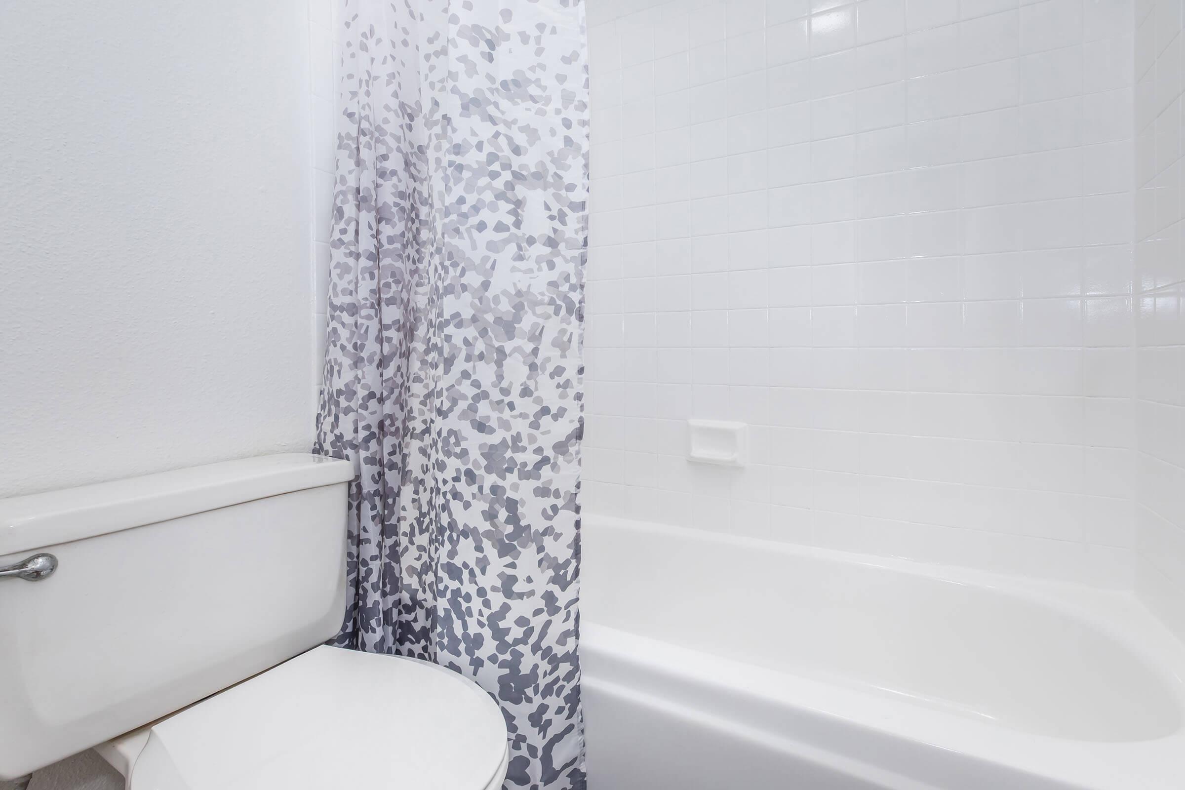 A clean bathroom featuring a white bathtub and a shower with a patterned curtain. There is a white toilet positioned next to the tub, and the walls are tiled in light colors, creating a fresh and modern atmosphere.