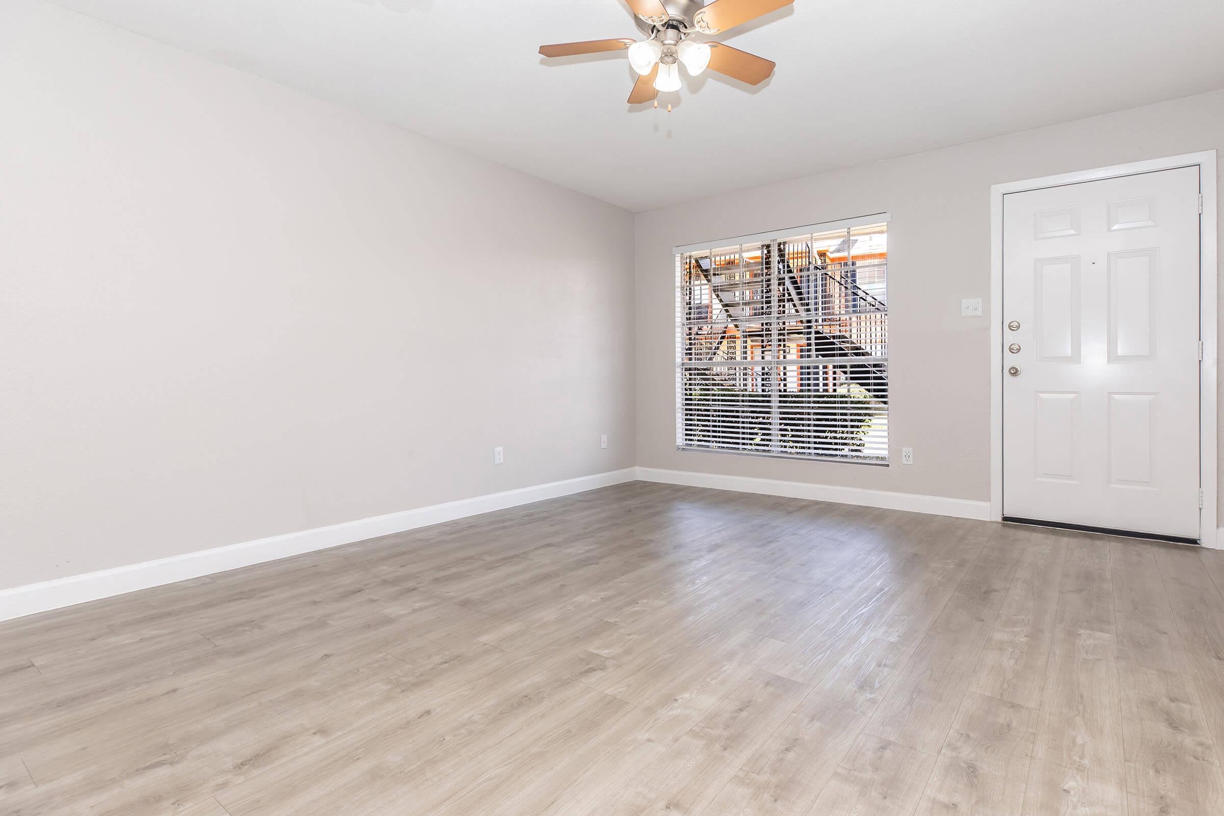 A spacious, empty living room featuring light-colored walls and a wood laminate floor. There is a ceiling fan with light fixtures and a large window with blinds allowing natural light. A doorway leads to the entrance of the room.