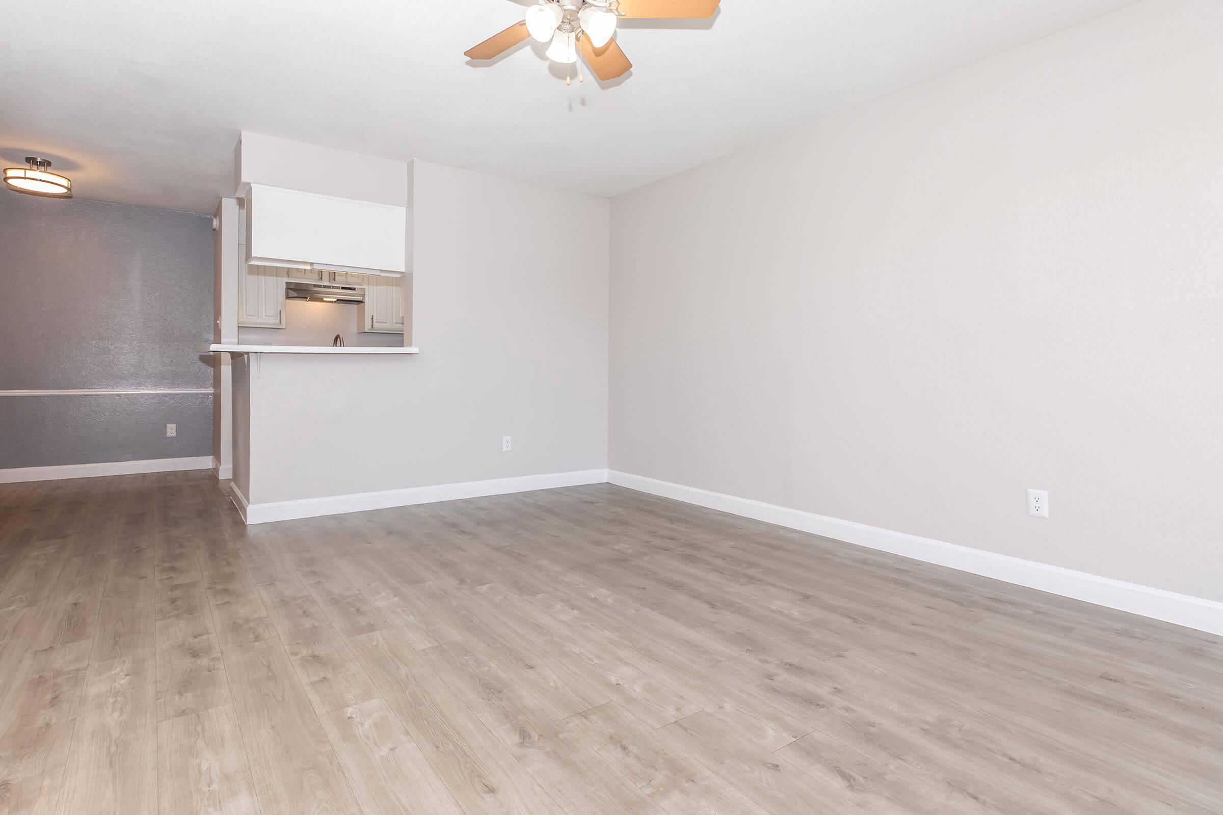 A spacious, empty room with light-colored walls and a ceiling fan. The floor is finished in a light wood look, and there is a small kitchen area visible in the background. Natural light enters the space, enhancing its airy feel. The overall atmosphere is clean and minimalistic, ideal for personalization.