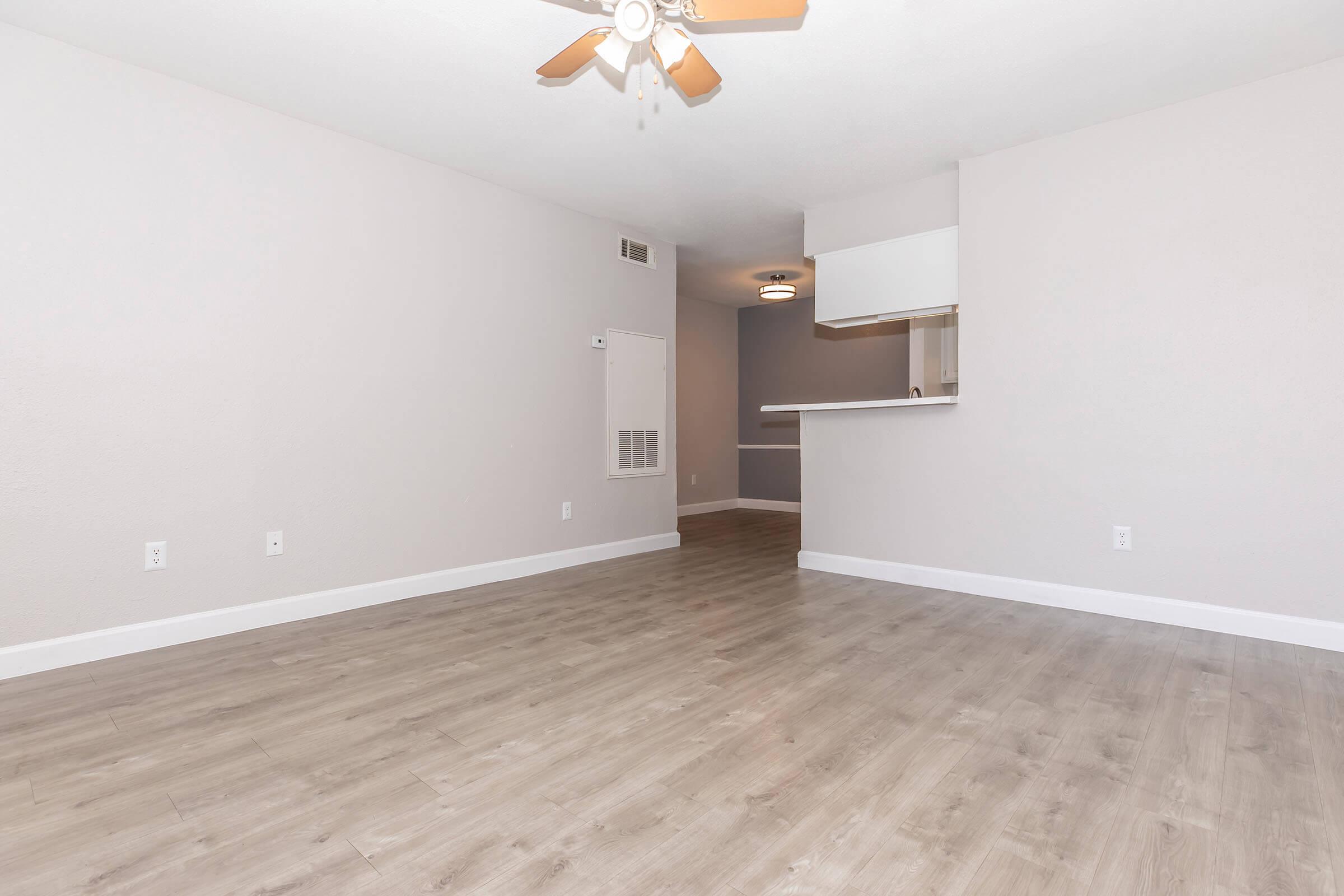 A spacious, light-filled room with light gray walls and a wooden floor. A ceiling fan is visible above. There is an open kitchen area in the background with a bar counter, creating a seamless flow between the living space and kitchen. The overall atmosphere is modern and inviting.