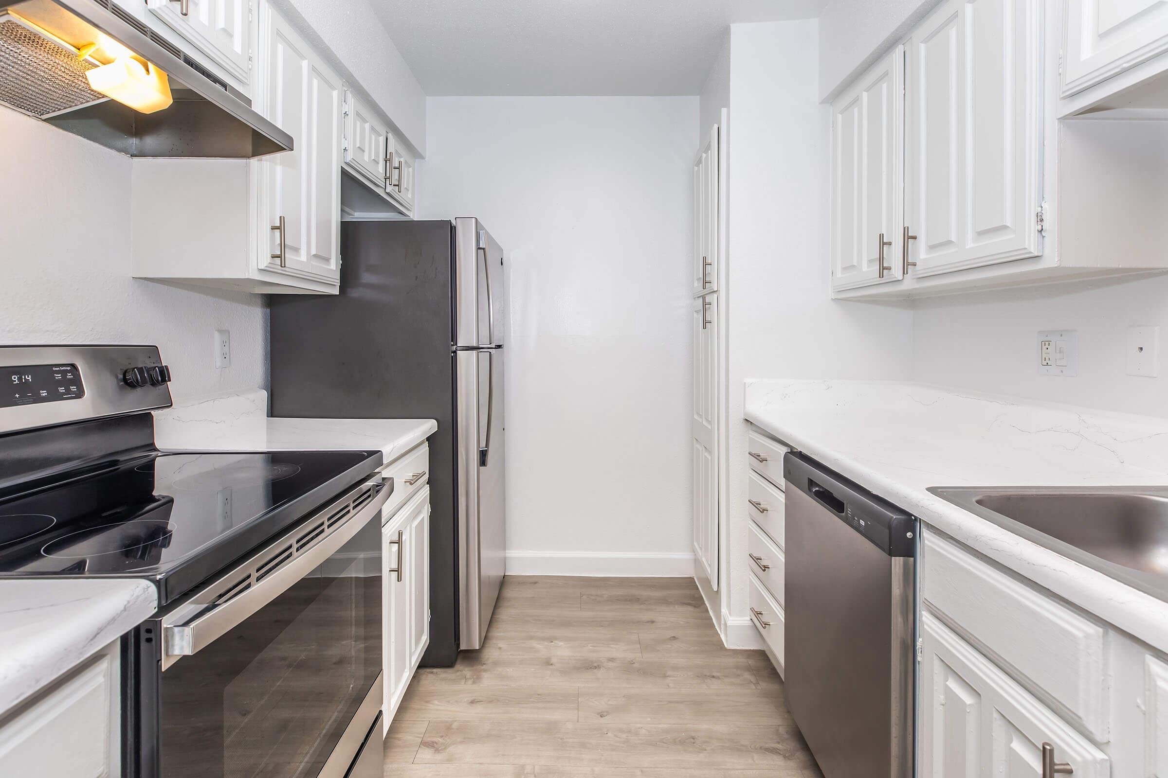 A modern kitchen with white cabinets, stainless steel appliances, including an oven, microwave, refrigerator, and dishwasher. The countertop is light-colored, and the flooring is laminate wood. The kitchen is well-lit with natural light and has a clean, minimalistic design.
