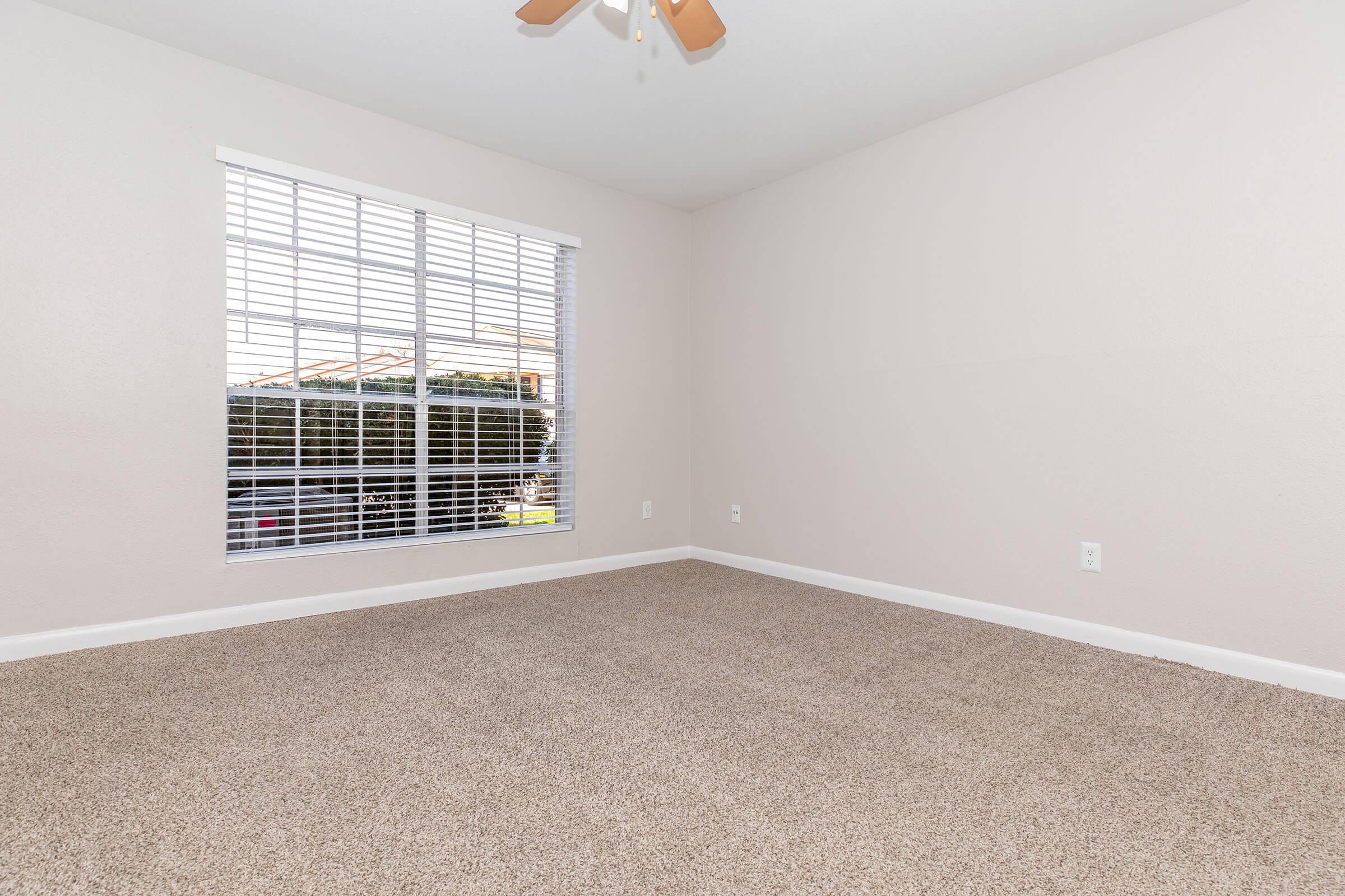 A vacant room with light beige walls and a soft carpeted floor. The room features a large window with white blinds, allowing natural light to enter. The ceiling has a light brown ceiling fan, contributing to a bright and airy atmosphere. The overall space is clean and uncluttered, ideal for various uses.