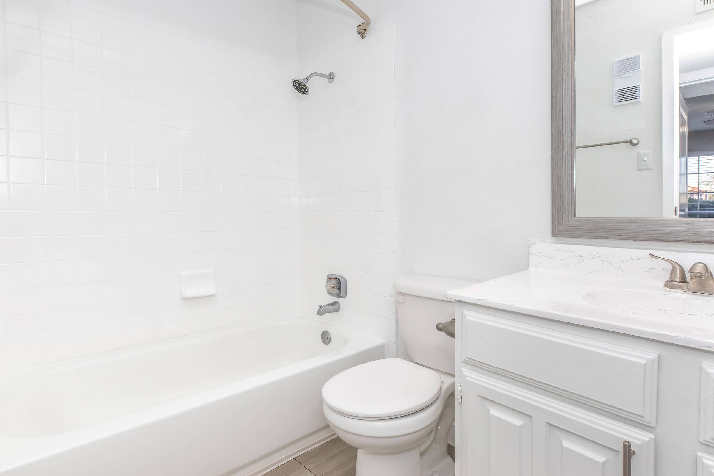 A clean and modern bathroom featuring a white tiled wall, a bathtub with a showerhead, a white toilet, and a sink with a marble countertop. A mirror above the sink reflects the space, and natural light can be seen coming from a window in the background.
