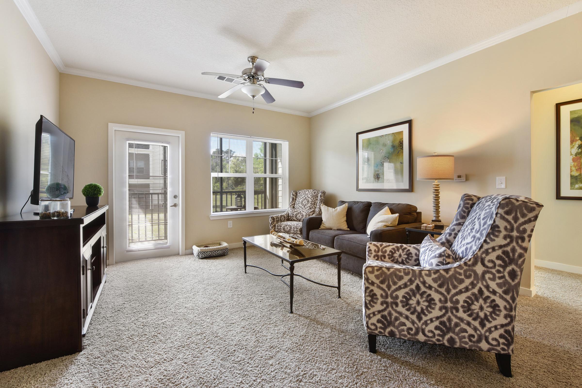 Cozy living room featuring a brown sofa, two patterned armchairs, and a decorative coffee table. A ceiling fan hangs above, and a flat-screen TV is mounted on a dark wooden cabinet. Natural light streams in through a window with a view of the outdoors, complementing the warm beige walls and plush carpet.