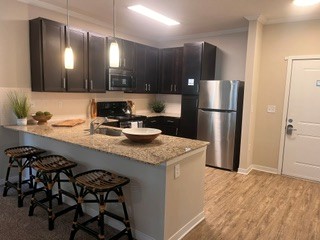 A modern kitchen featuring dark wood cabinets, granite countertops, and stainless steel appliances. There are three woven stools at the kitchen island, with decorative bowls and plants on the counter. Natural light illuminates the space, and a door to the right leads outside.