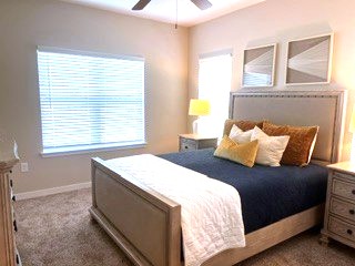A cozy bedroom featuring a plush bed with blue and white bedding, adorned with decorative pillows in shades of yellow and gold. Natural light filters through a window with blinds, and a bedside lamp casts a warm glow. Two framed artworks hang above the bed, contributing to the room's inviting atmosphere.