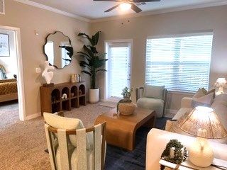 A cozy living room featuring a light-colored sofa, two accent chairs, and a wooden coffee table. There’s a modern lamp on the side table, a decorative mirror on the wall, and large windows allowing natural light. Potted plants add greenery to the space, enhancing the inviting atmosphere.