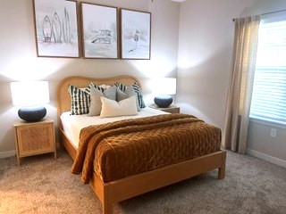 A cozy bedroom featuring a neatly made bed with a brown throw blanket and decorative pillows, two bedside lamps, and modern artwork on the walls. The room has light beige walls, a warm carpet, and a window with sheer curtains allowing natural light to enter.