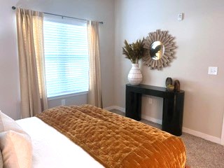A cozy bedroom featuring a bed with a textured orange blanket, a modern black console table, and a decorative mirror. Natural light streams in through white curtains, enhancing the warm tones of the decor. A vase with dried plants adds a touch of greenery to the room.