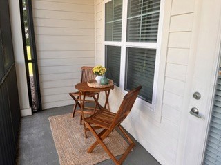 A cozy porch area featuring a small round table with a floral centerpiece and two wooden folding chairs. The setting includes light-colored walls and large windows with white blinds, creating a welcoming atmosphere. A textured rug adds warmth to the space, complemented by a view of greenery outside.
