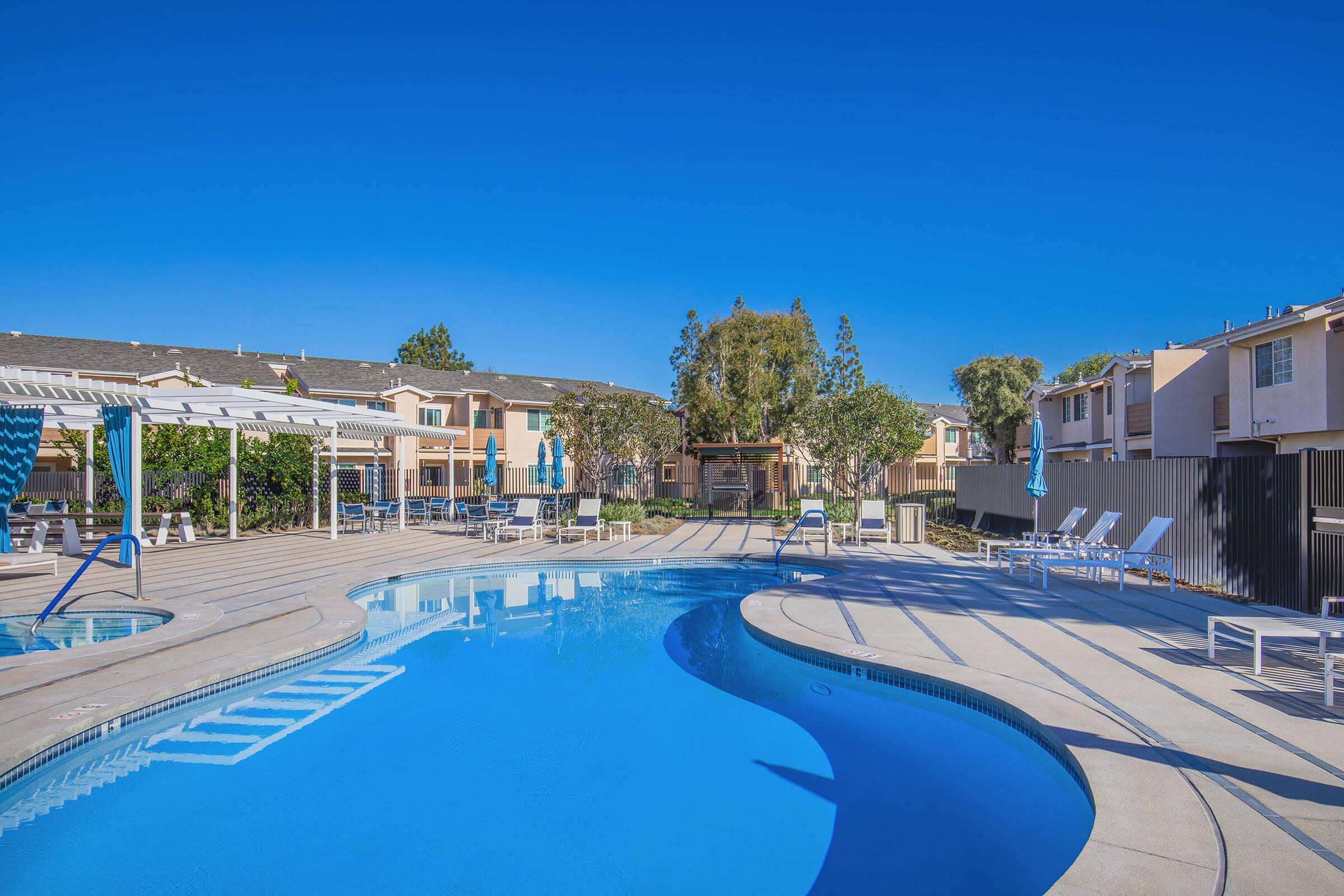 A sunny outdoor swimming pool area surrounded by palm trees and lounge chairs. The pool features a curvy design with steps leading in, and nearby are shaded seating areas with umbrellas. Residential buildings can be seen in the background under a clear blue sky.