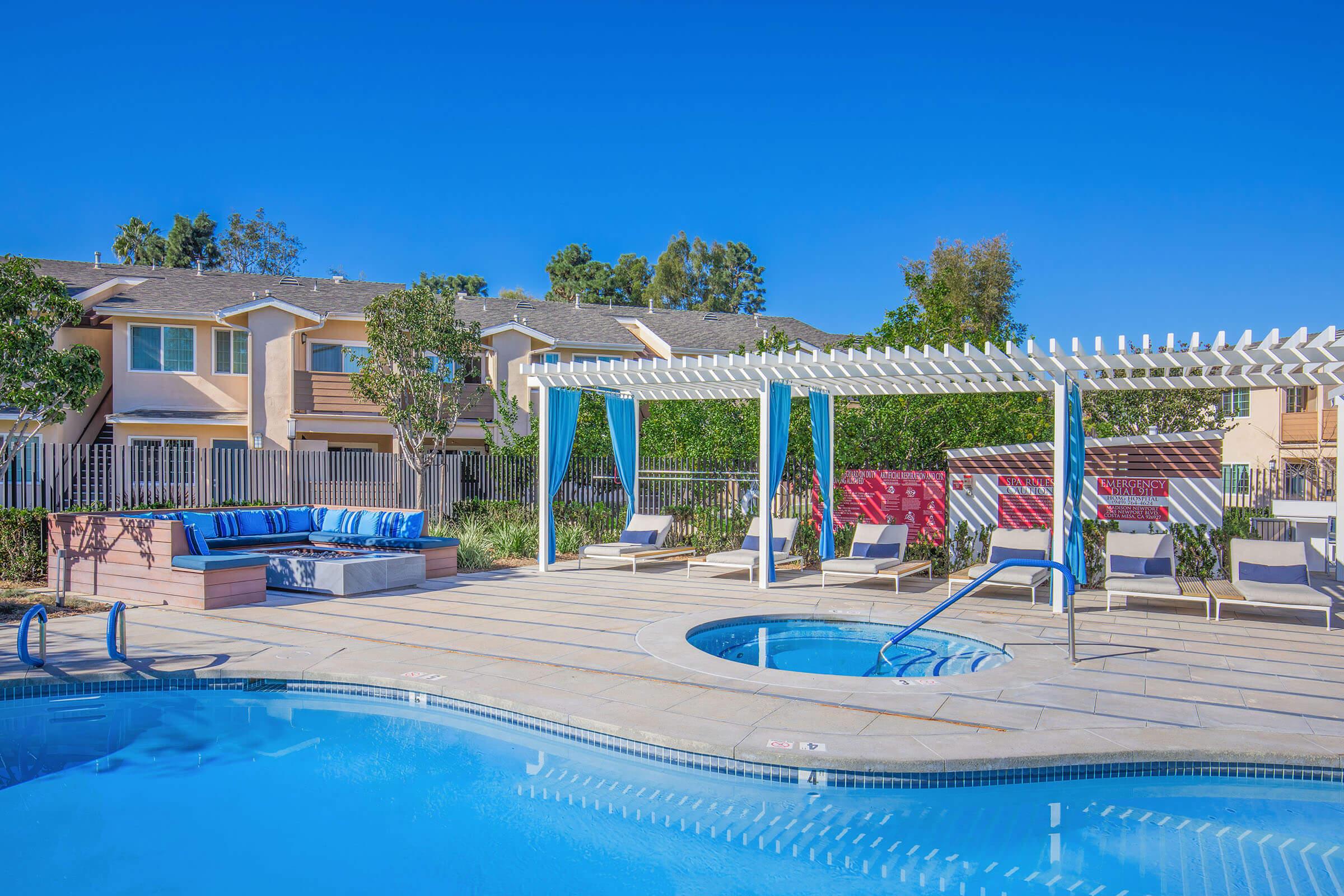 A sunny poolside area featuring a sparkling blue pool and a hot tub. Surrounding the pool are lounge chairs and a shaded seating area with a sofa under a pergola. Lush green trees and residential buildings are visible in the background, creating a relaxing outdoor space.