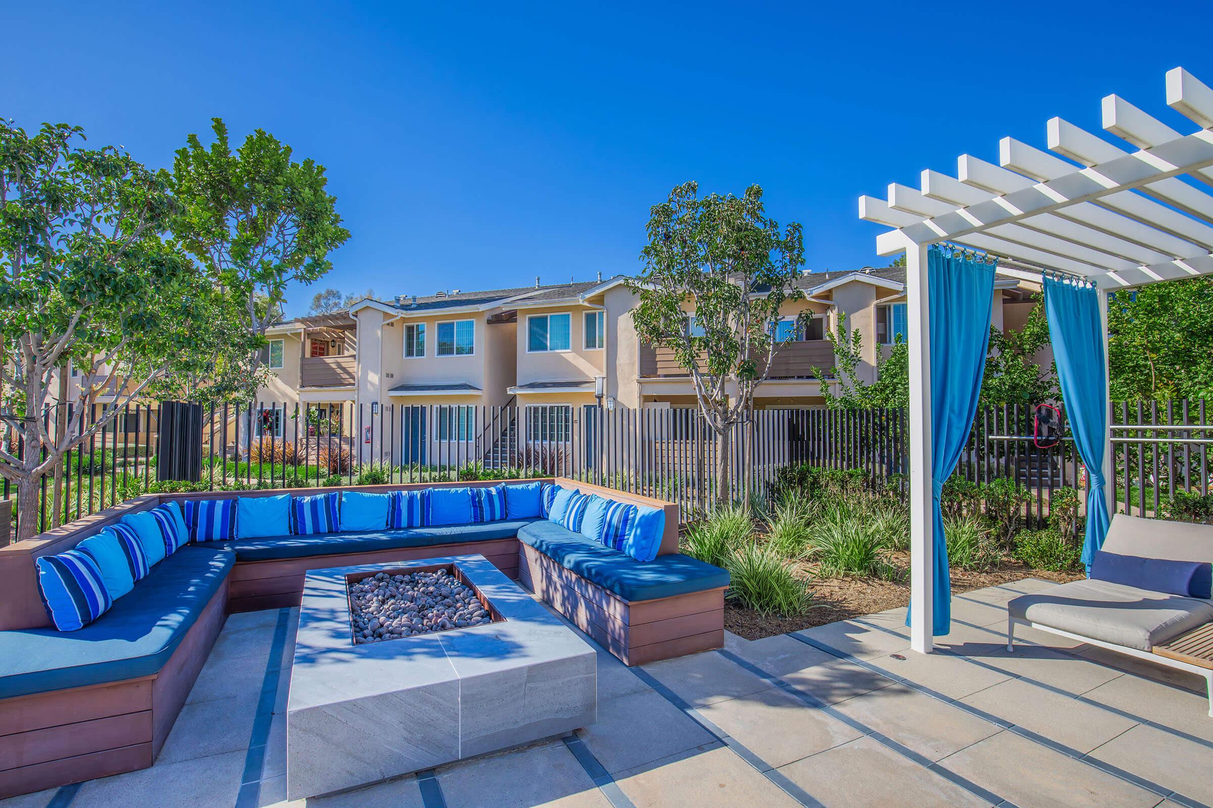 A landscaped outdoor lounge area featuring a curved seating arrangement with blue and white cushions surrounding a stone fire pit. Nearby, there are two trees, a pergola with flowing blue curtains, and several modern residential buildings in the background under a clear blue sky.